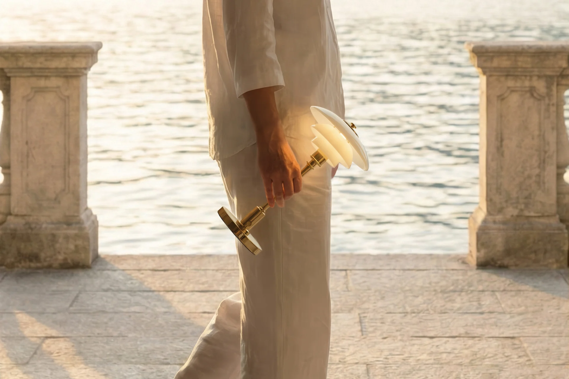 Persona de blanco con lámpara iluminada de latón y pantallas blancas, camina junto a agua brillante y pilares de piedra.