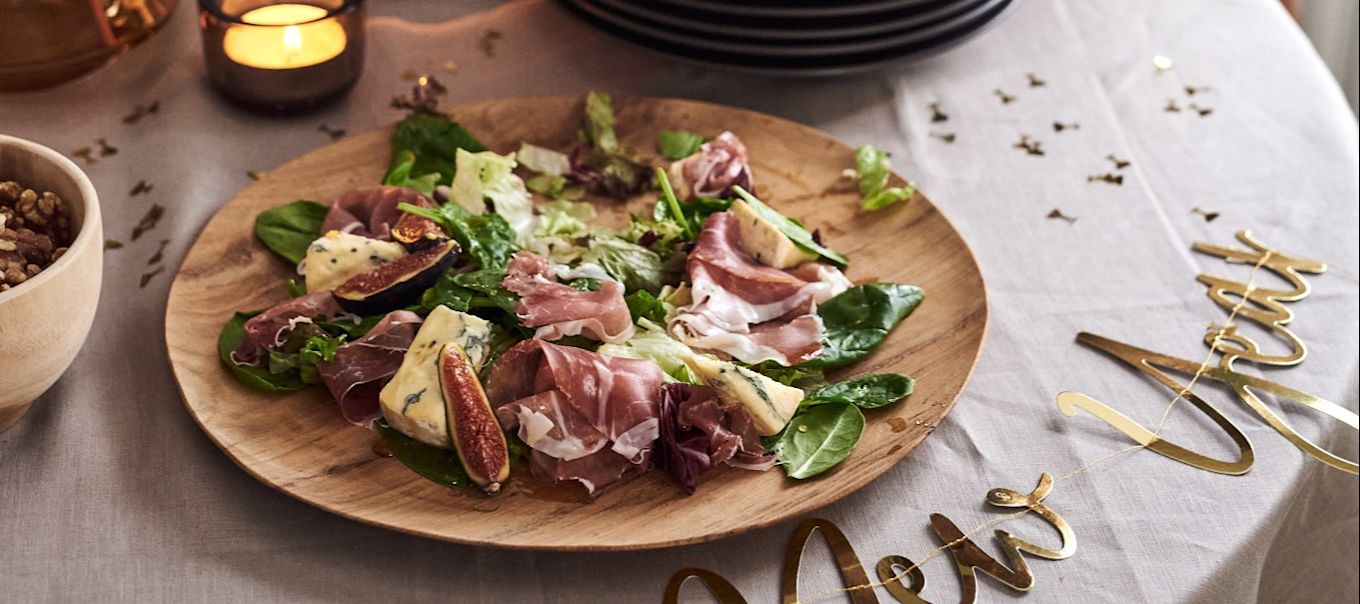 Te proponemos 5 sencillas recetas de entrantes para tu fiesta de fin de año. Una deliciosa ensalada de jamón serrano y queso azul junto a un cartel de Feliz Año Nuevo.