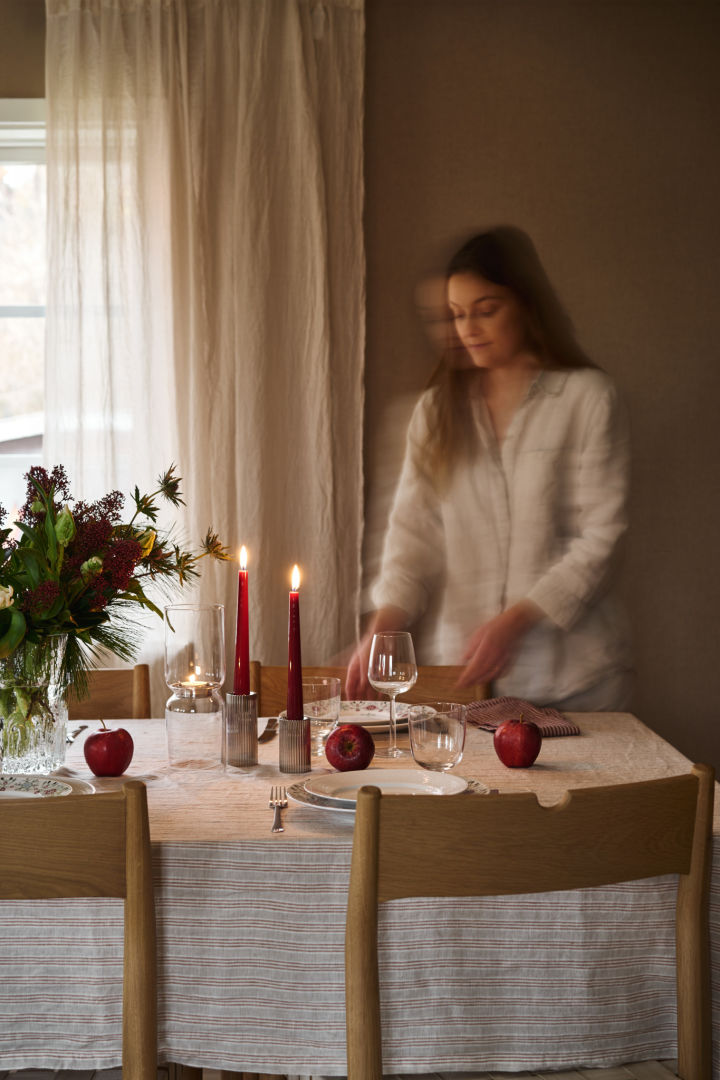 Una mujer pone una mesa navideña con velas rojas, porcelana navideña estampada y un sencillo mantel de rayas.