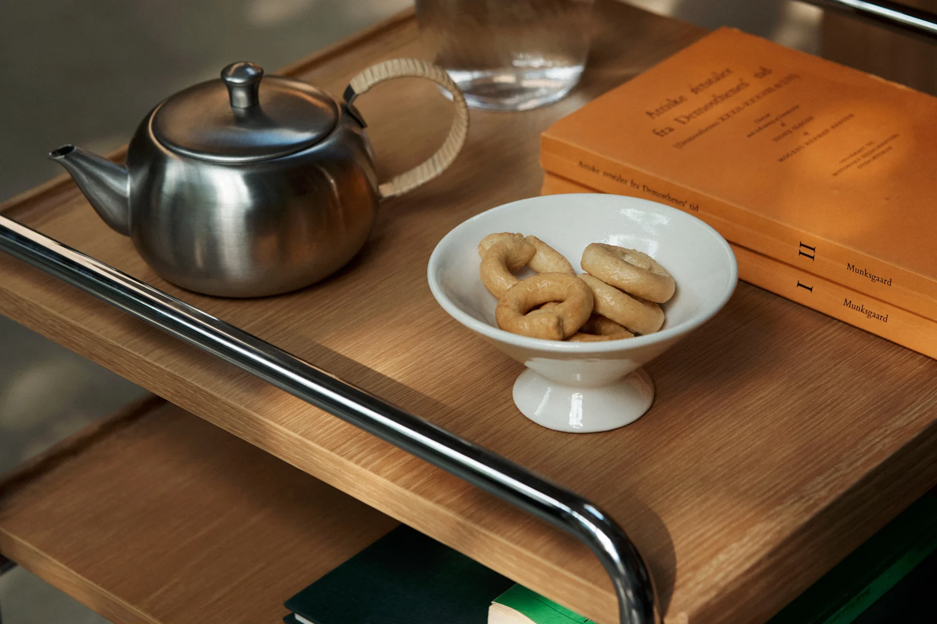 Una tetera de acero inoxidable, un cuenco blanco con galletas de aro y dos libros naranjas en un estante de madera.