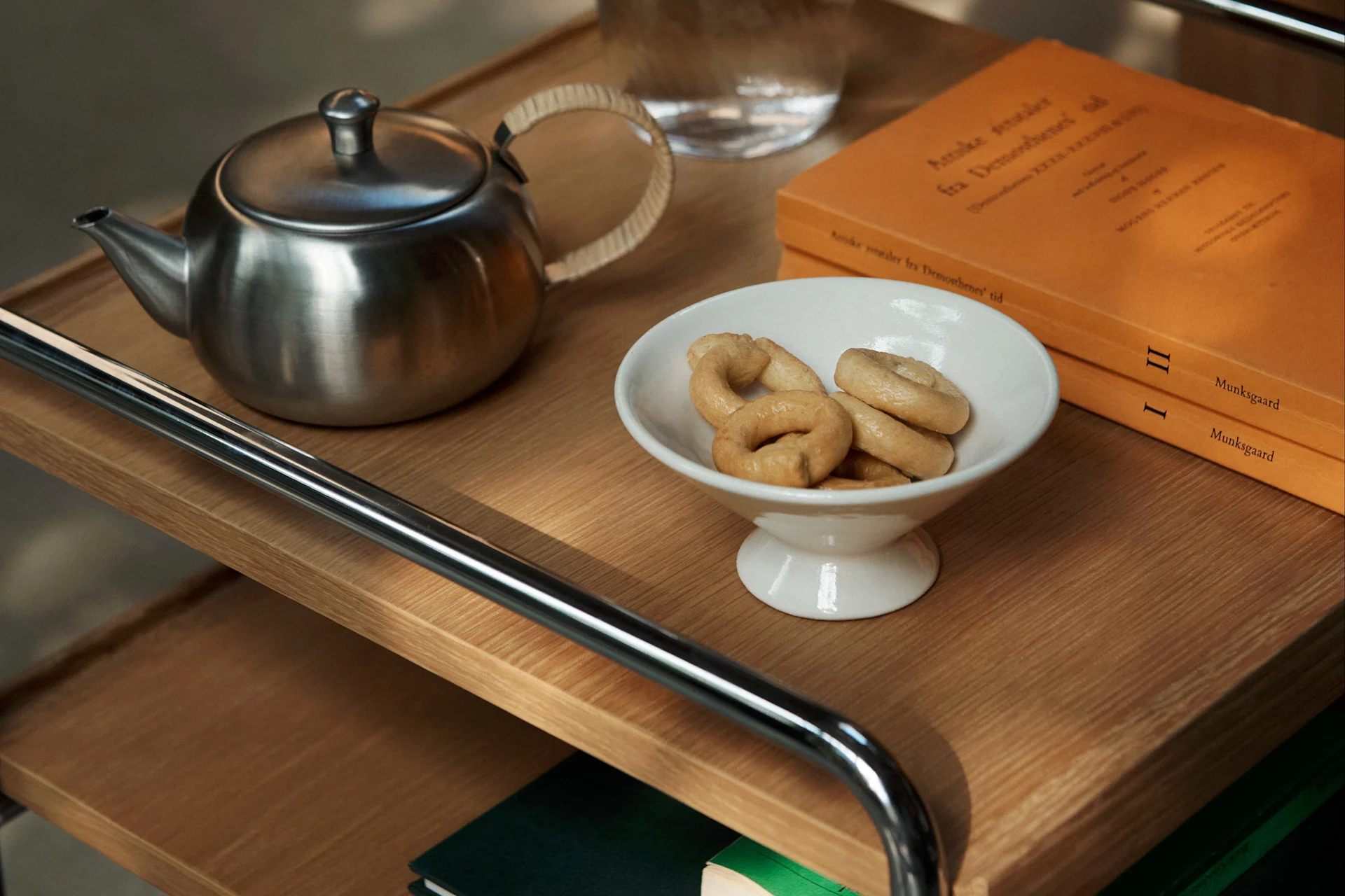 Una tetera de metal, un cuenco blanco con galletas en forma de anillo y dos libros naranjas en un carrito de madera.