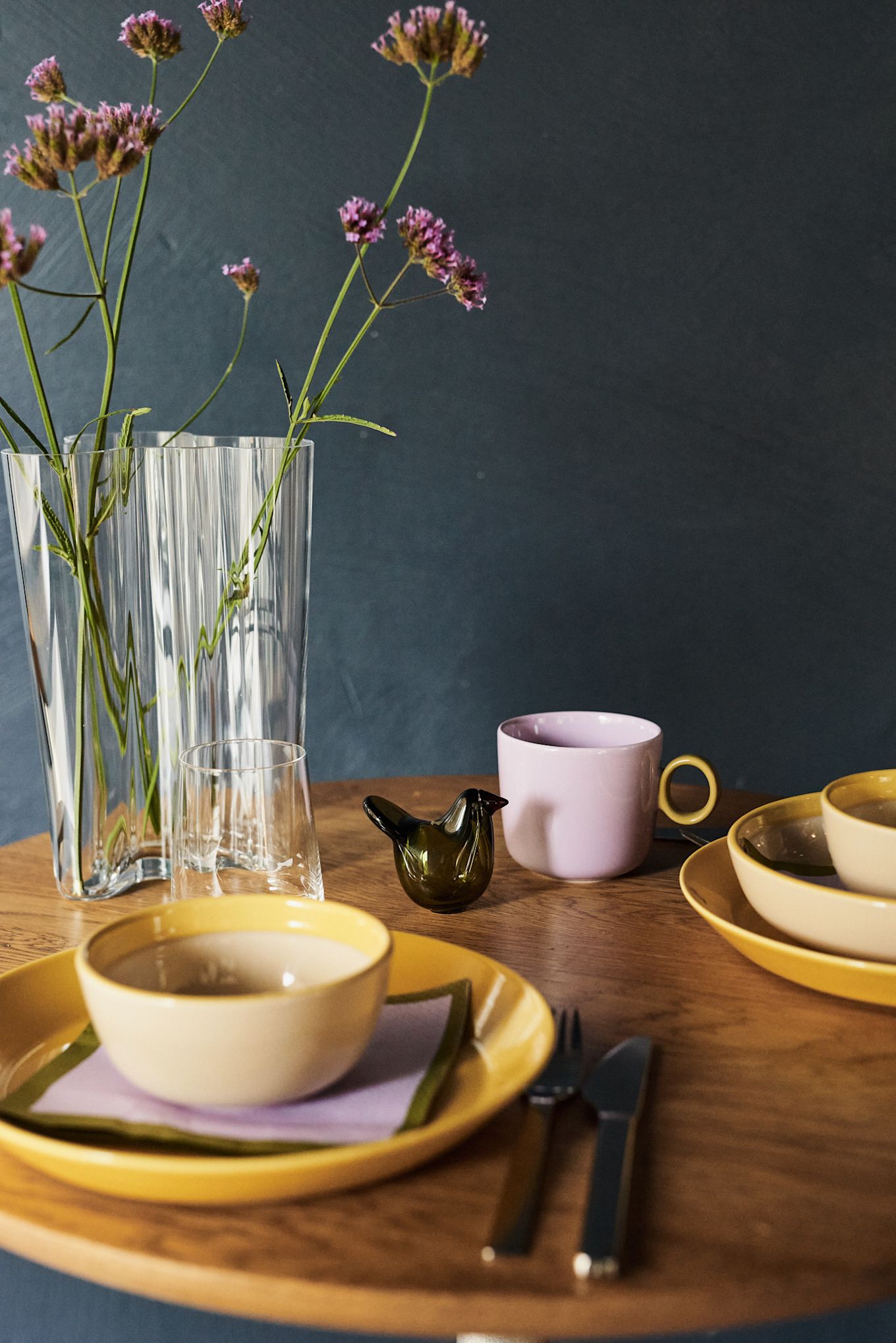 Aquí ves una idea de decoración de mesa en amarillo con tazas moradas en contraste y un pájaro de vidrio verde musgo en un ambiente de estilo retro de cafetería.