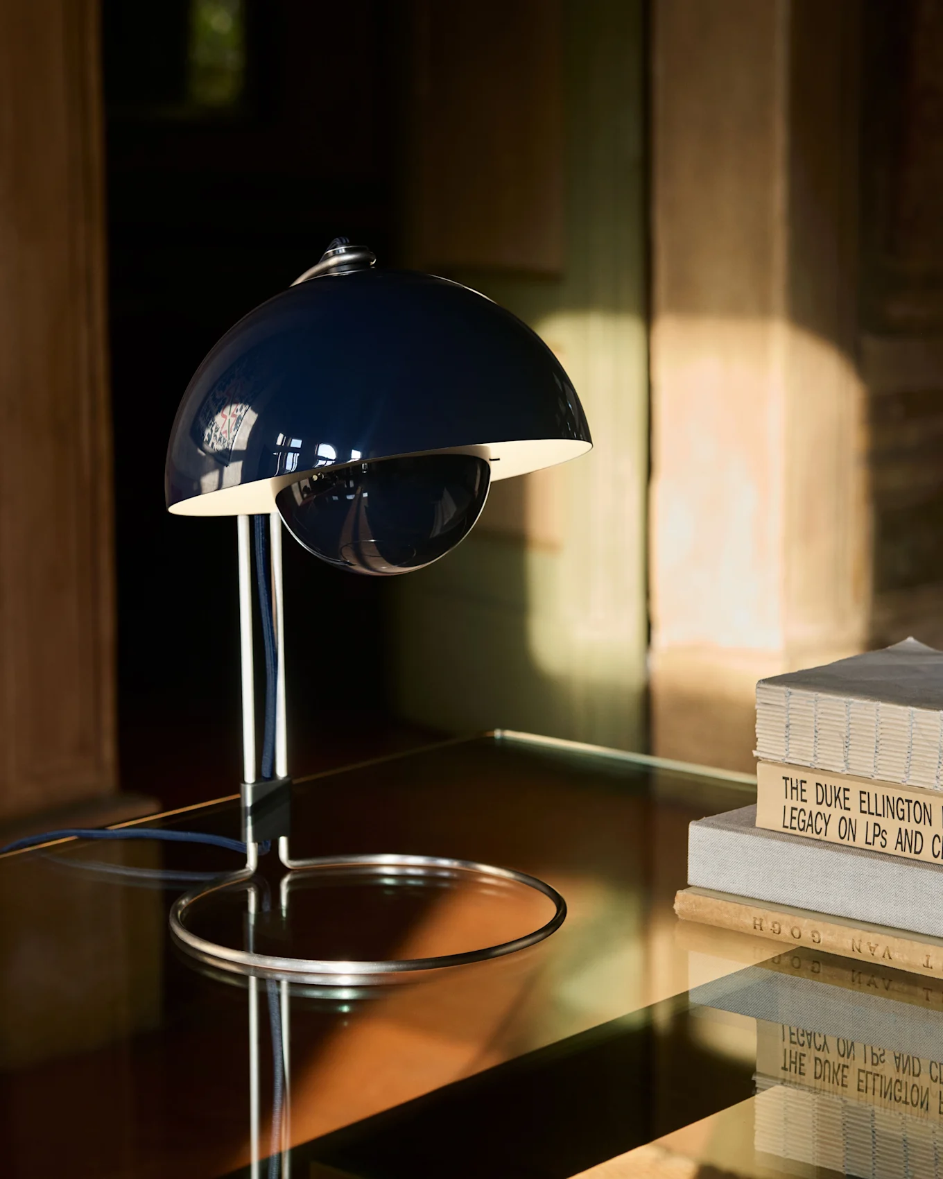 Una lámpara de mesa azul oscuro con forma de campana y base cromada sobre una mesa reflectante junto a una pila de libros.
