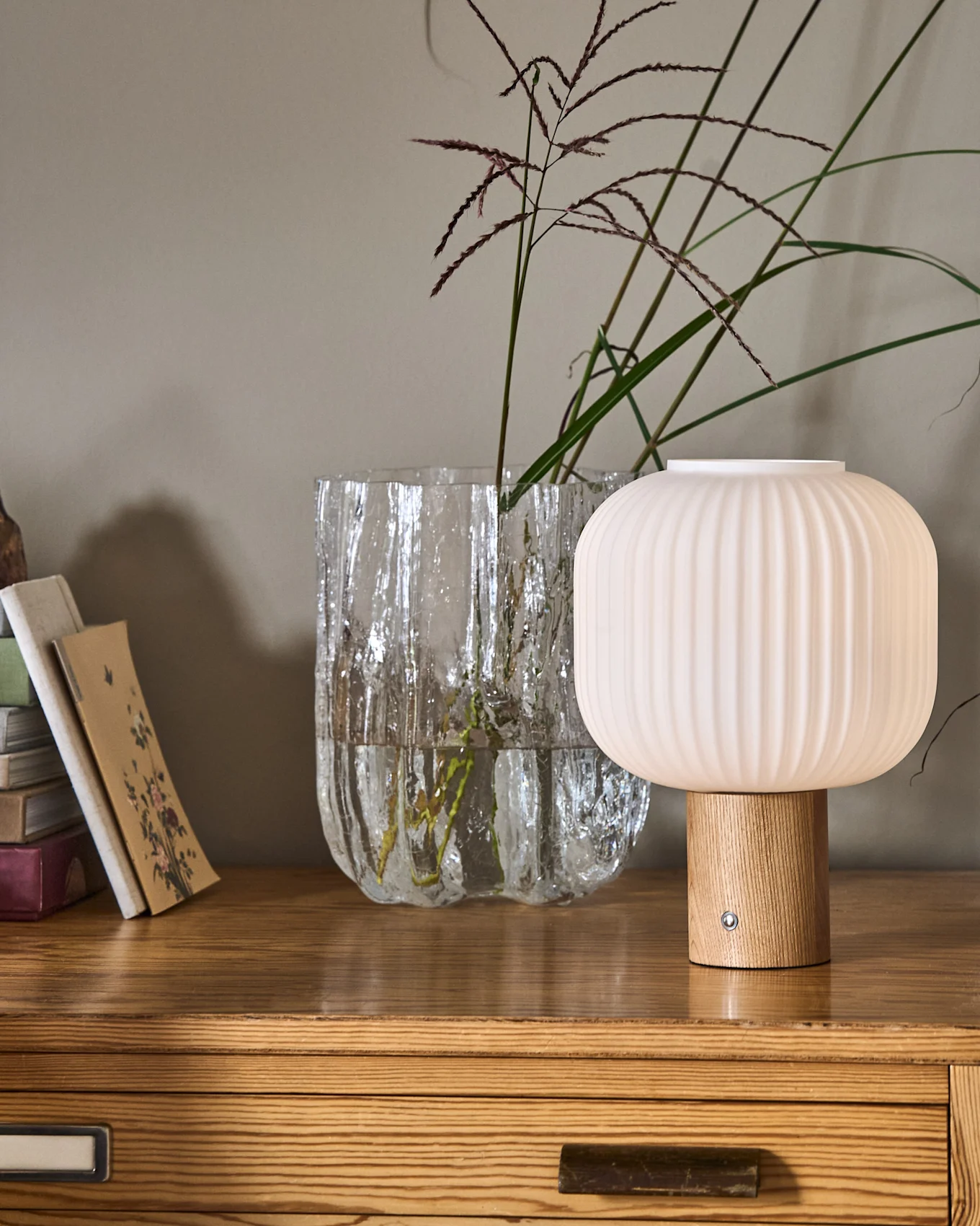 Una lámpara acanalada en forma de hongo con base de madera junto a un jarrón de cristal texturizado con plantas y una pila de libros sobre una cómoda de madera.