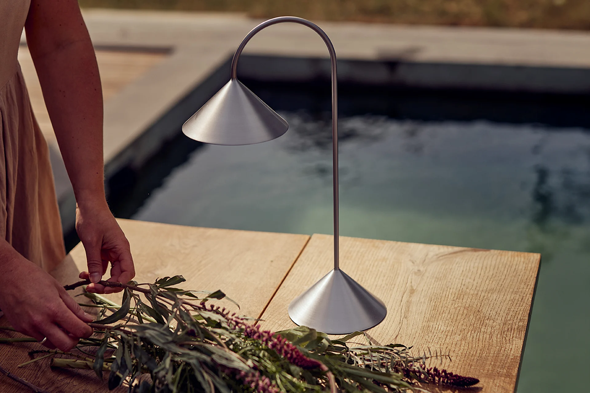 Manos arreglando flores en una mesa de madera junto a una lámpara de mesa de metal cepillado junto a una piscina.