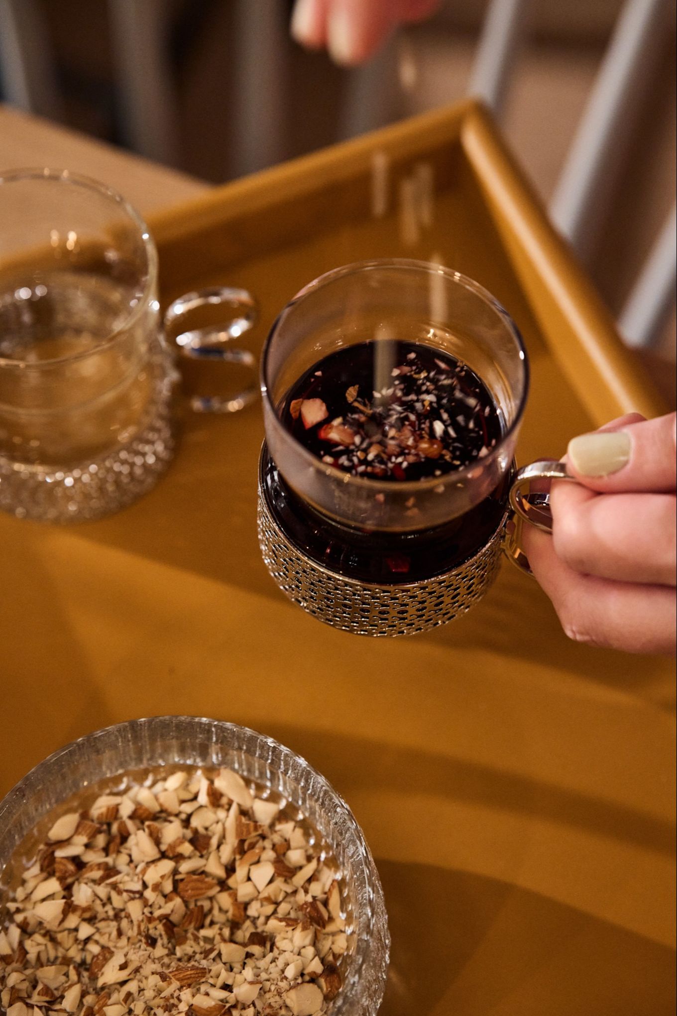 Una mano espolvorea almendras en un vaso tradicional de glögg sobre una mesa navideña nórdica.