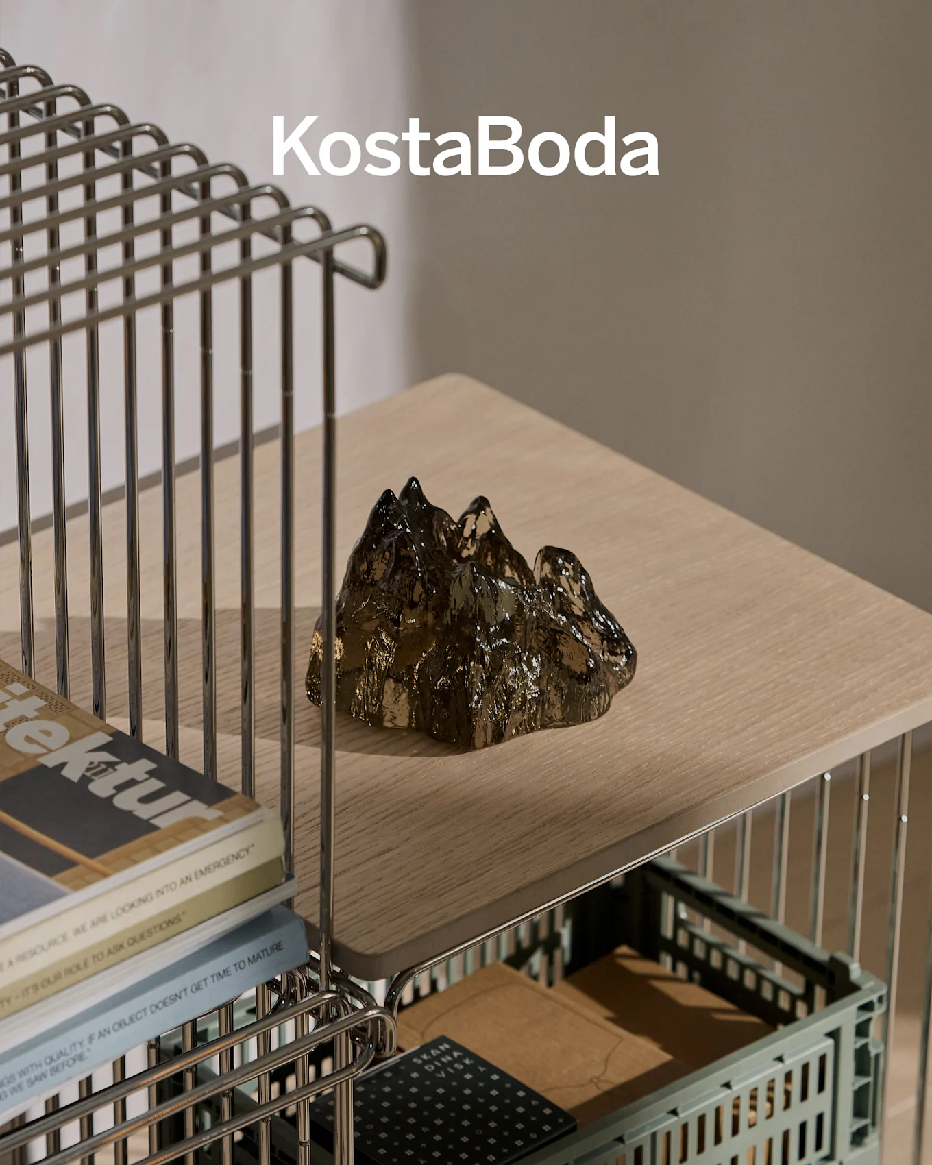 Marca KostaBoda sobre una escultura de vidrio oscura, transparente y con forma de montaña en un estante de madera clara.