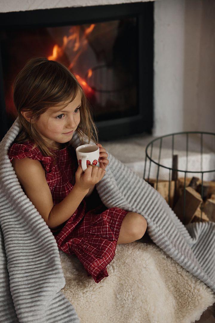 Las celebraciones navideñas suecas también incluyen una taza de chocolate caliente o glögg. Aquí puedes ver a una niña con la tazay la manta polar, ambas de Design House Stockholm, sentada en un sofá frente a la chimenea.