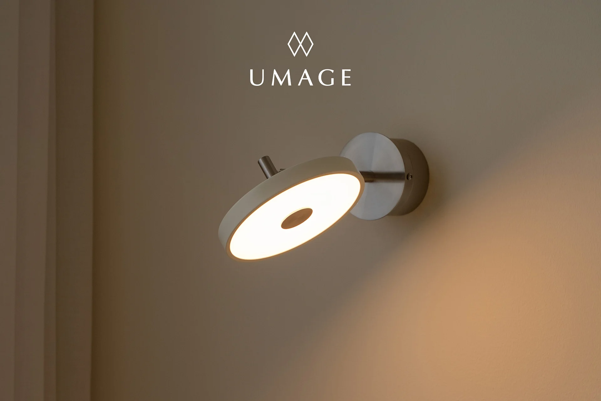 Una lámpara de pared circular moderna con luz cálida, montada en una pared marrón claro. Logo UMA arriba.