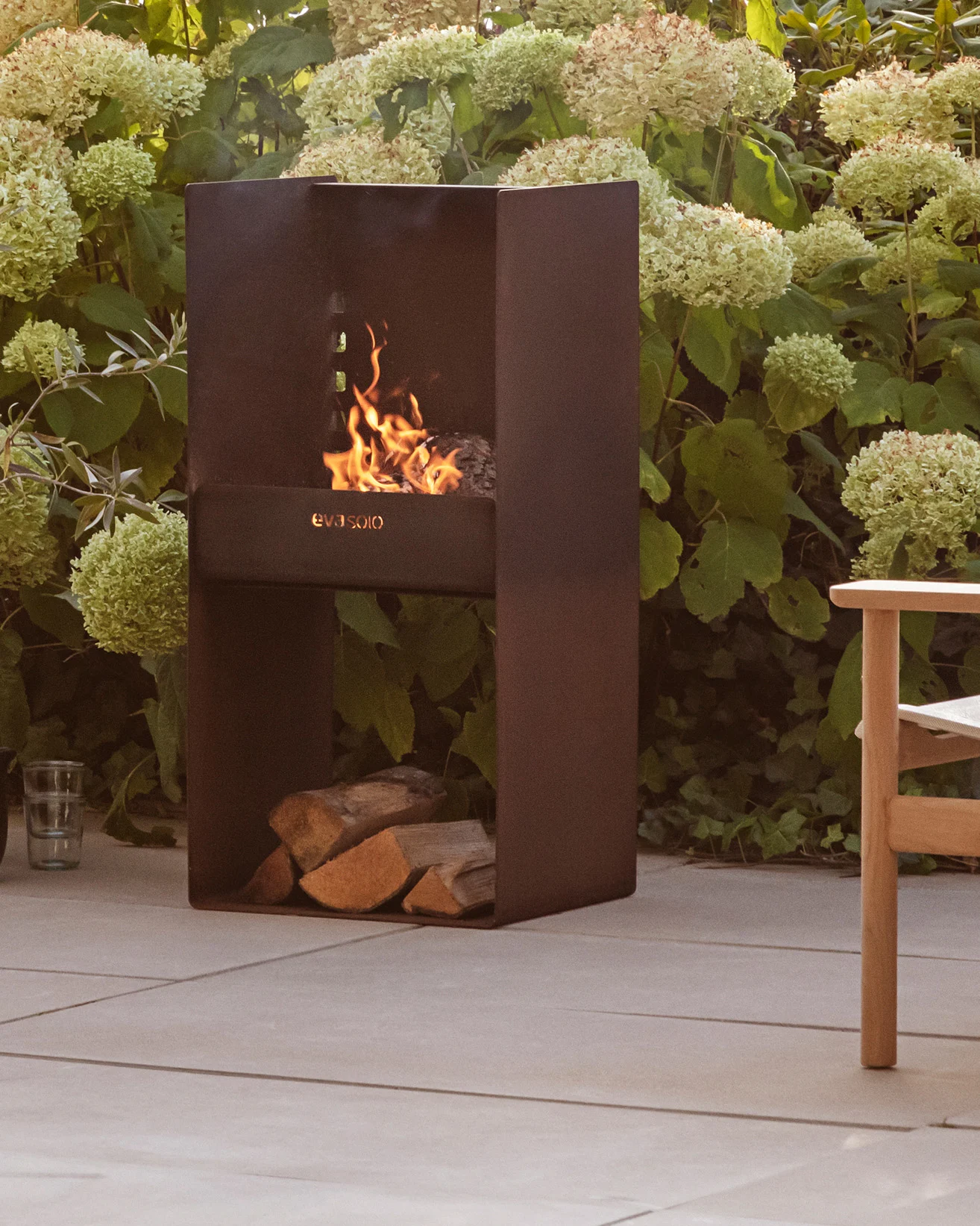 Moderna chimenea exterior de metal oscuro con fuego encendido, leña apilada debajo, rodeada de plantas verdes y un patio.