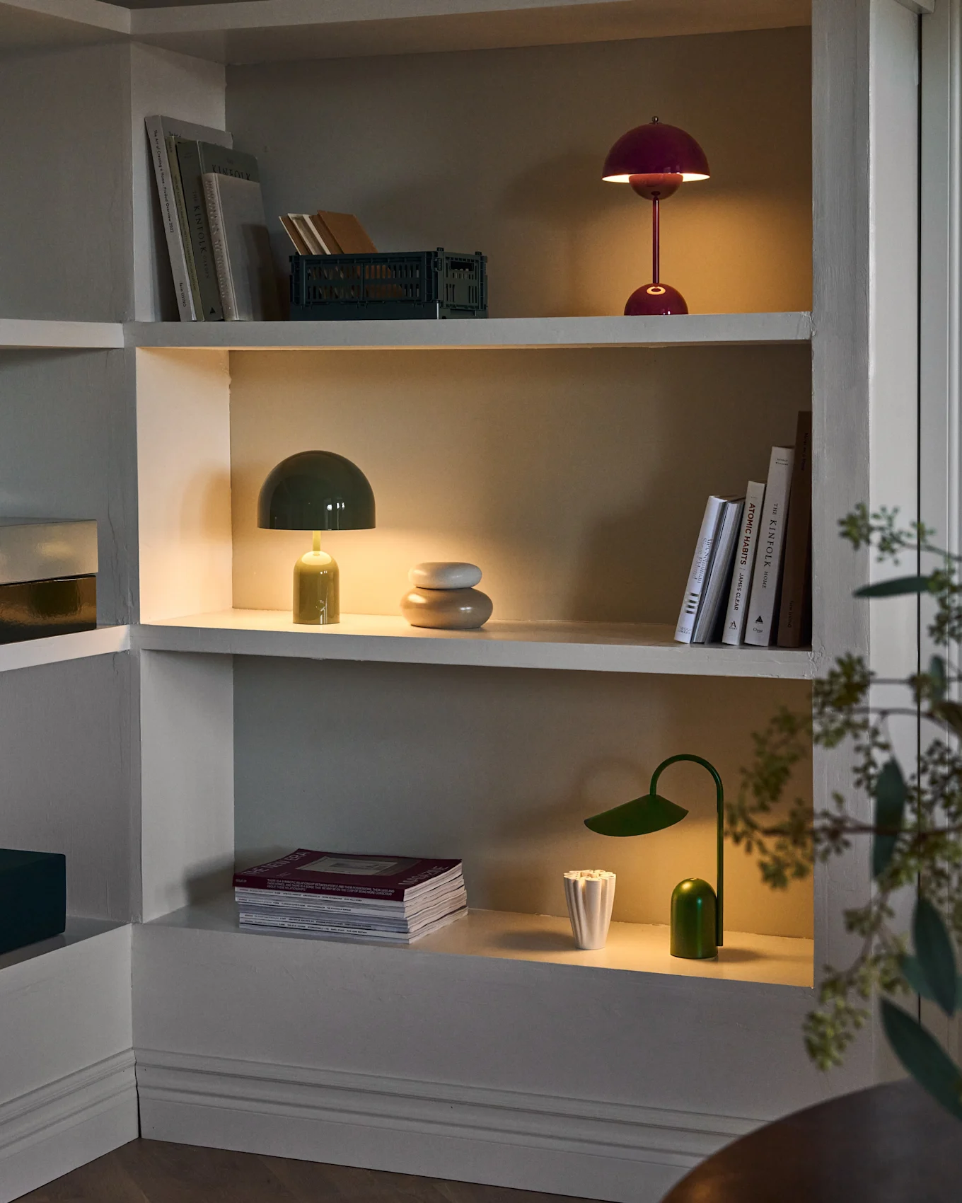 Estantes iluminados en mueble empotrado, con lámparas de seta rojas y verdes, lámpara de escritorio verde, libros y decoración.