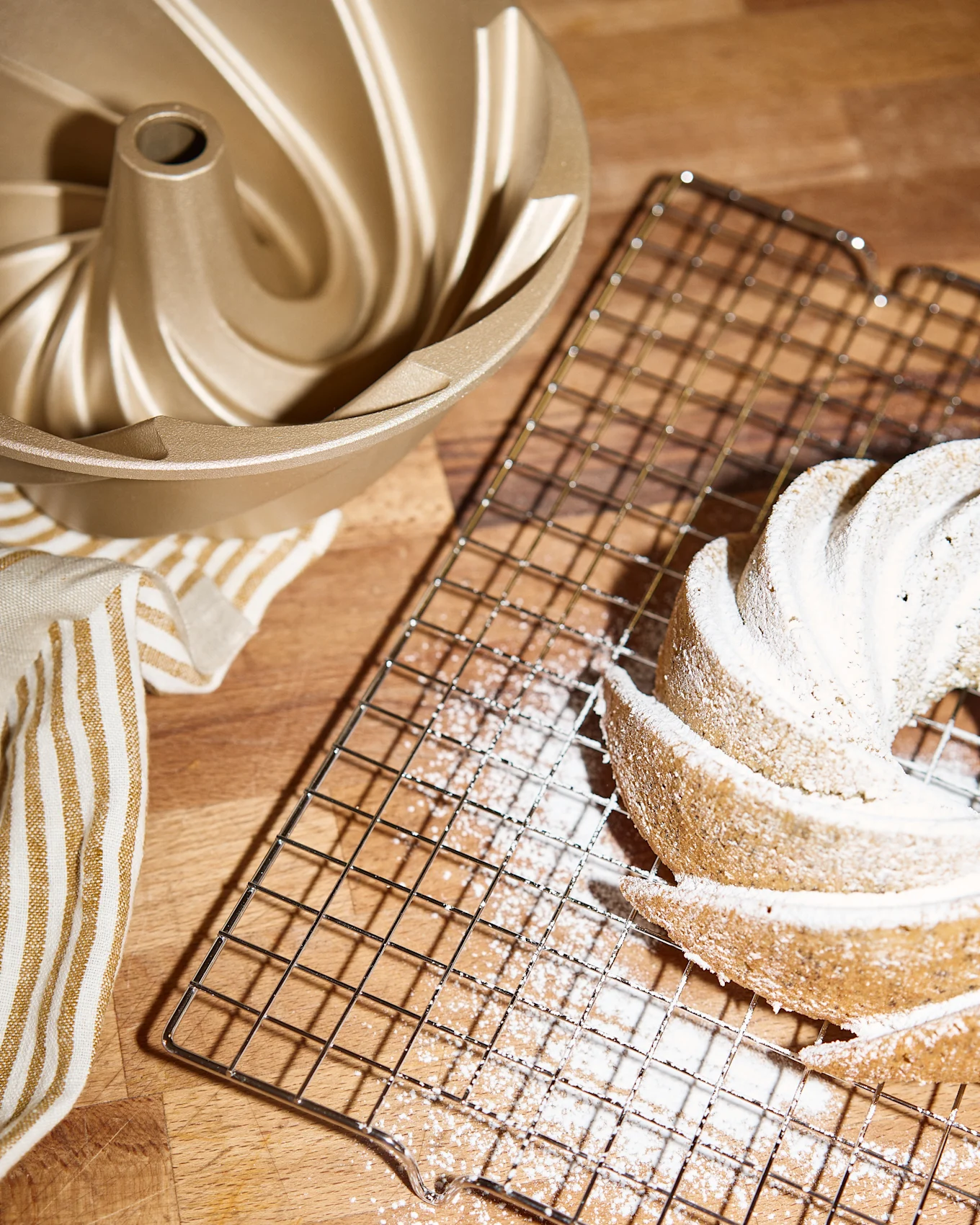 Un molde dorado para bundt cake, una rejilla de enfriamiento con un bundt cake espolvoreado con azúcar glas y una toalla rayada sobre una superficie de madera.