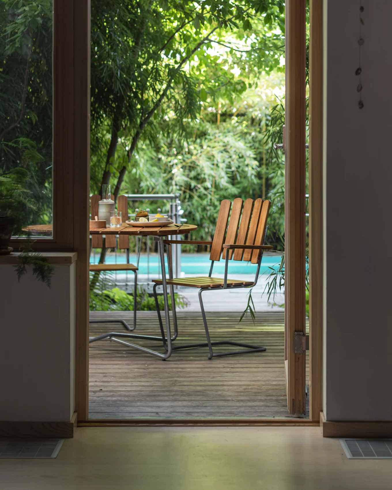 Vista a través de una puerta abierta a un patio exterior con una mesa de madera, sillas y una piscina rodeada de plantas verdes.