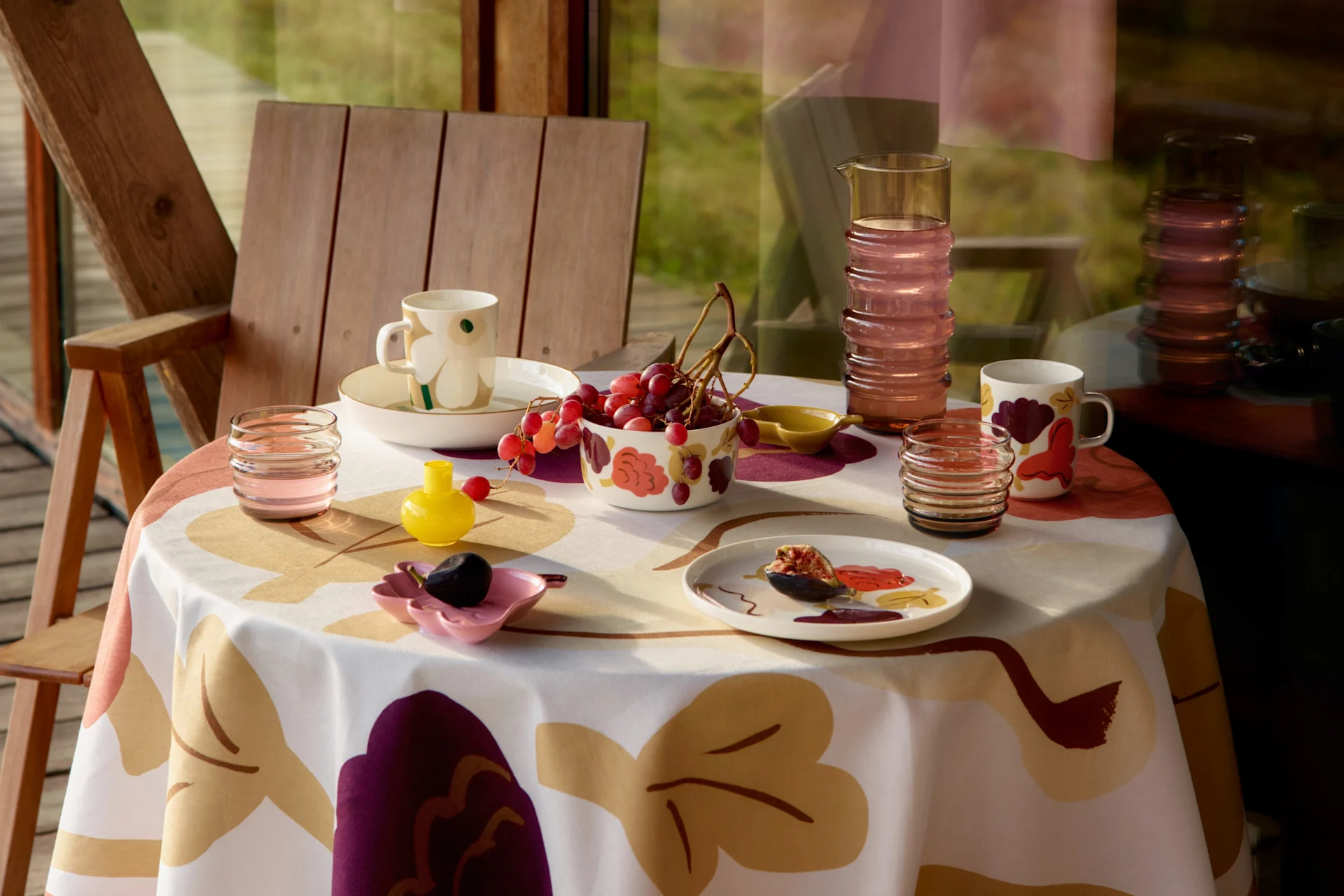 Una mesa puesta con un mantel estampado, tazas, cuencos con uvas, un higo en un plato, una jarra y vasos, junto a una silla de madera.