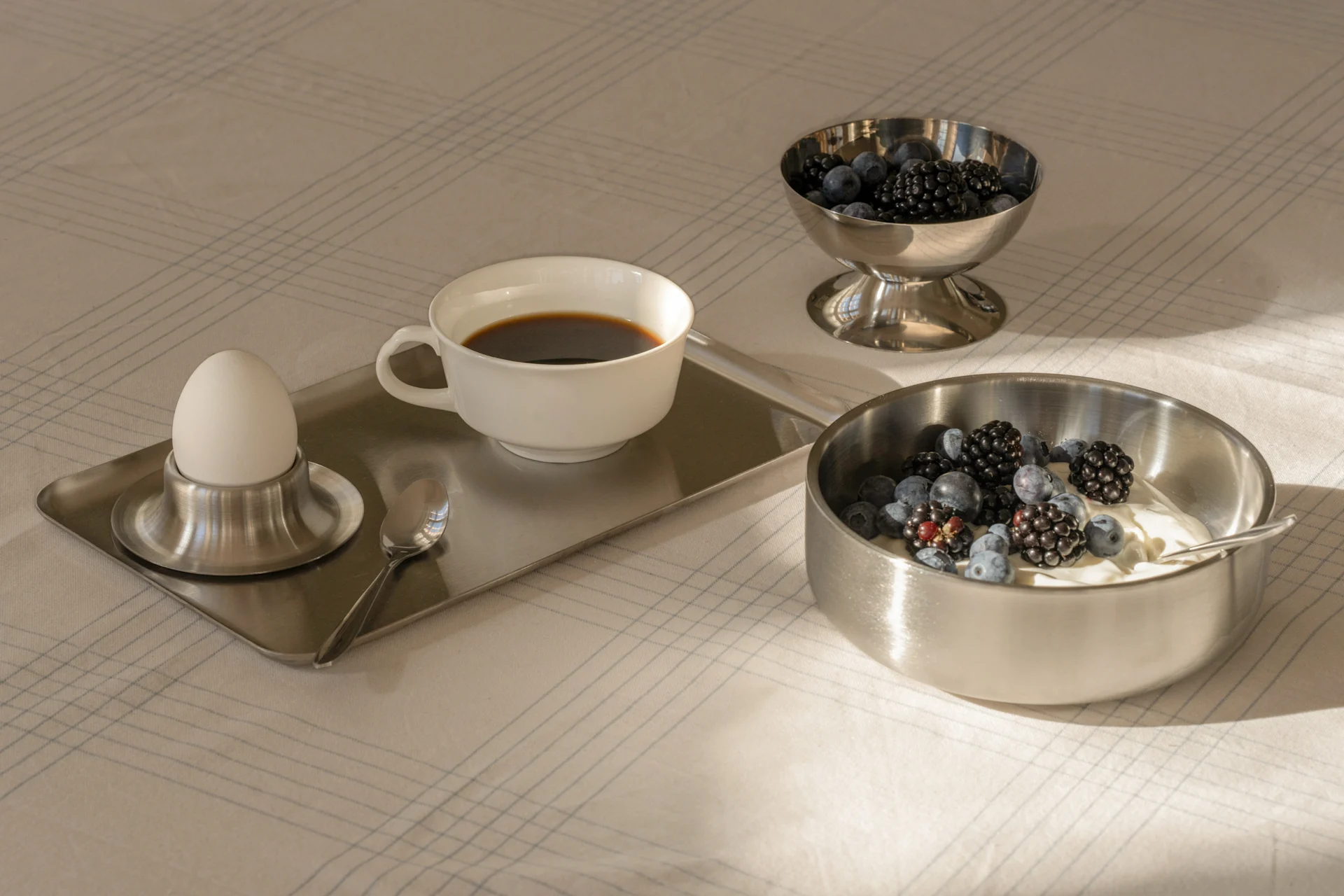 Desayuno con café, huevo cocido, yogur y bayas frescas en cuencos de plata sobre un mantel estampado.