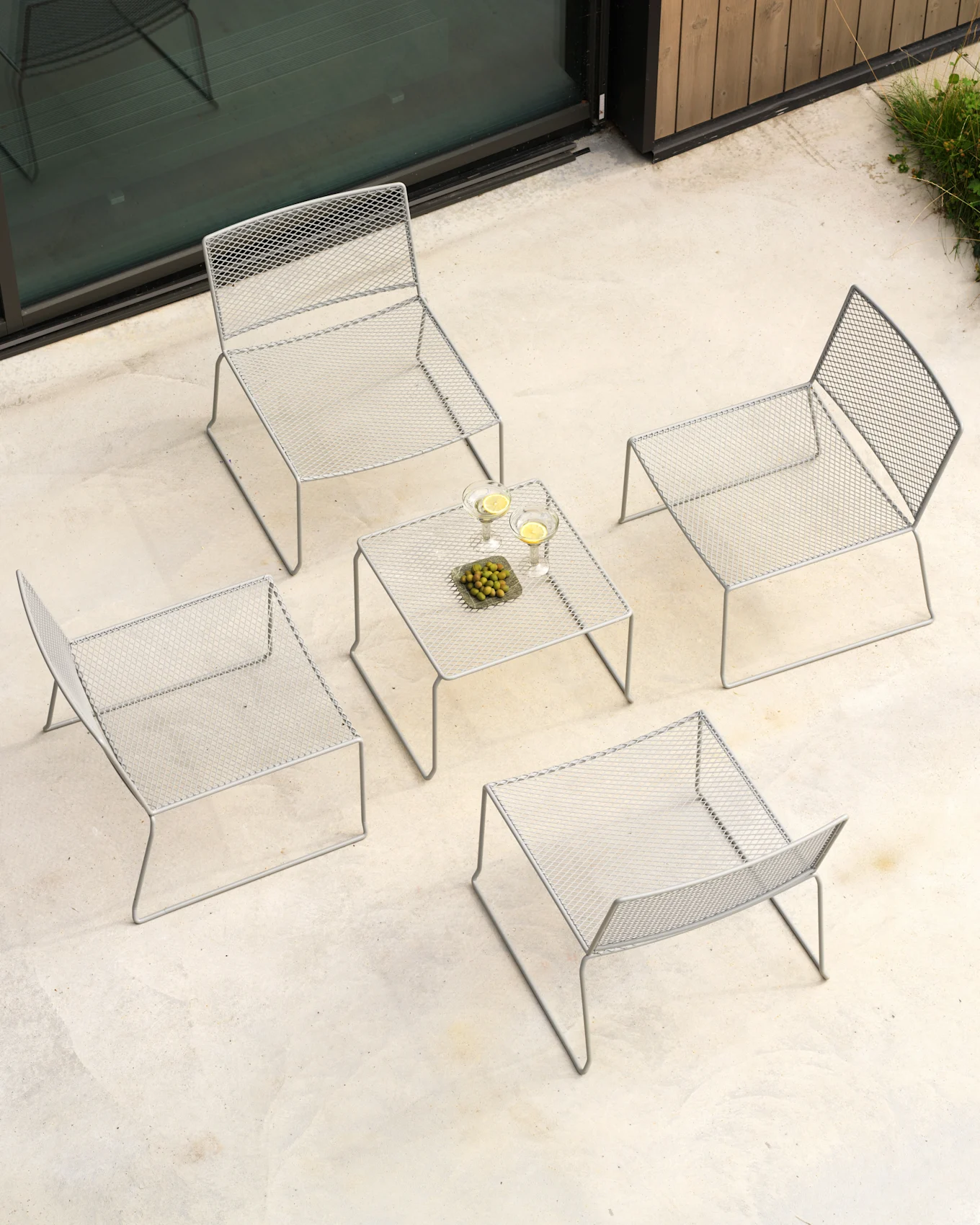 Vista aérea de un conjunto de patio exterior con cuatro sillas de malla metálica gris y una mesa de centro a juego con bebidas y aceitunas.