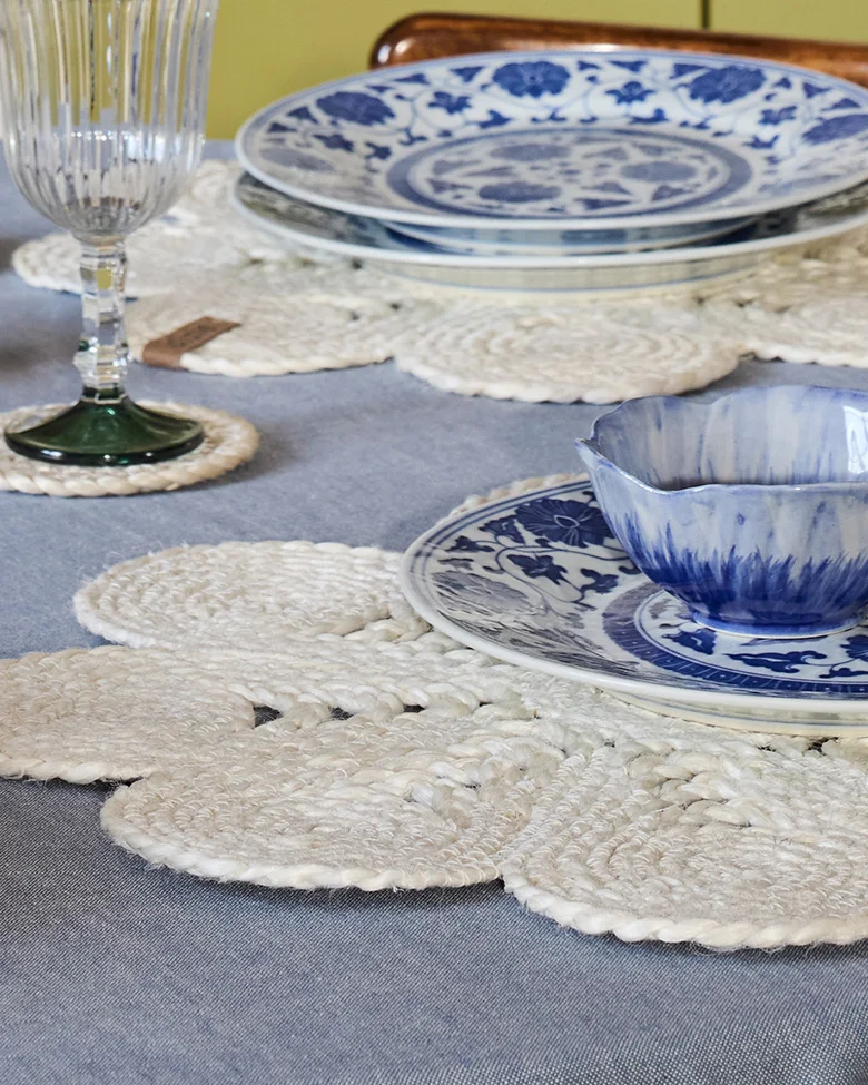 Mesa puesta con vajilla azul y blanca, manteles individuales florales trenzados blancos y un vaso estriado.