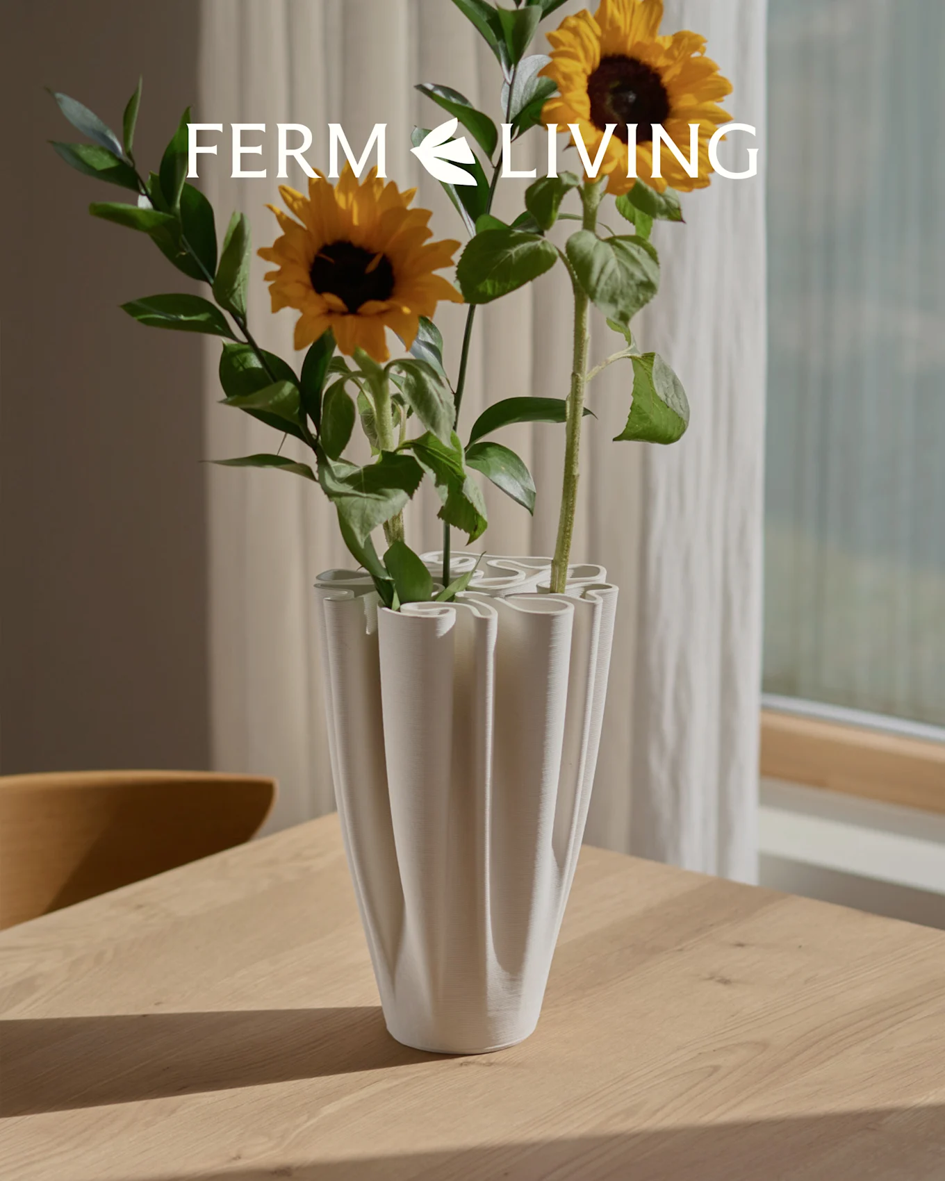 Girasoles en un jarrón blanco, estriado y ondulado sobre una mesa de madera. Superposición de logo Ferm Living.