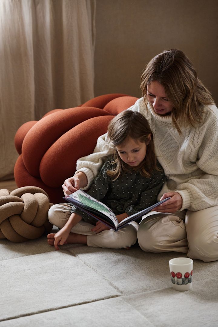 La lectura en voz alta de un cuento de Navidad sueco es parte integrante de las celebraciones navideñas en familia. Aquí vemos a una madre y a su hija sentadas en una alfombra hojeando un libro, rodeadas de un cojín Knot pequeño y otro grande, todos de Design House Stockholm.