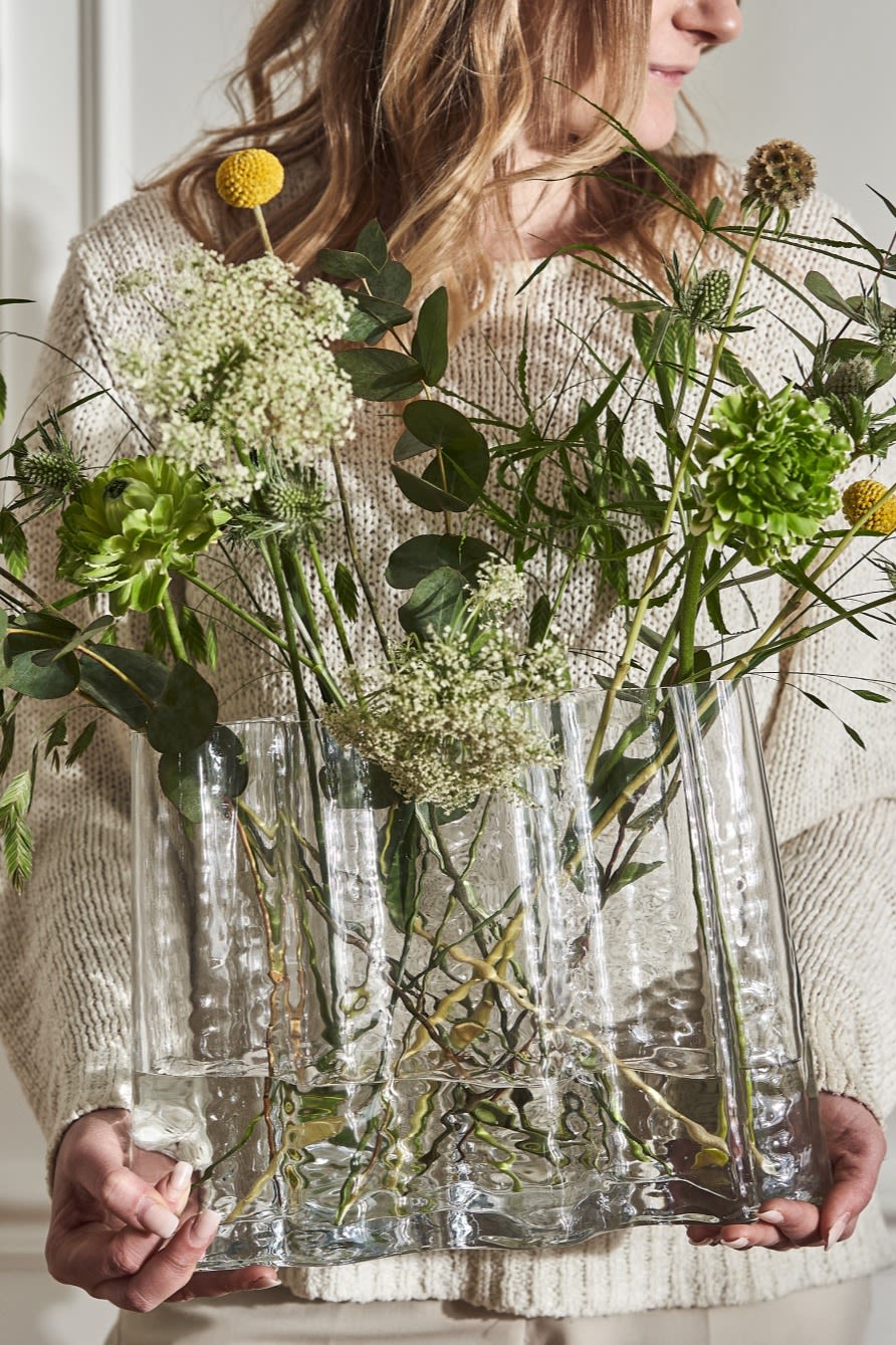 Una mujer vestida de crema sostiene el jarrón Gry lleno de flores silvestres de primavera. 