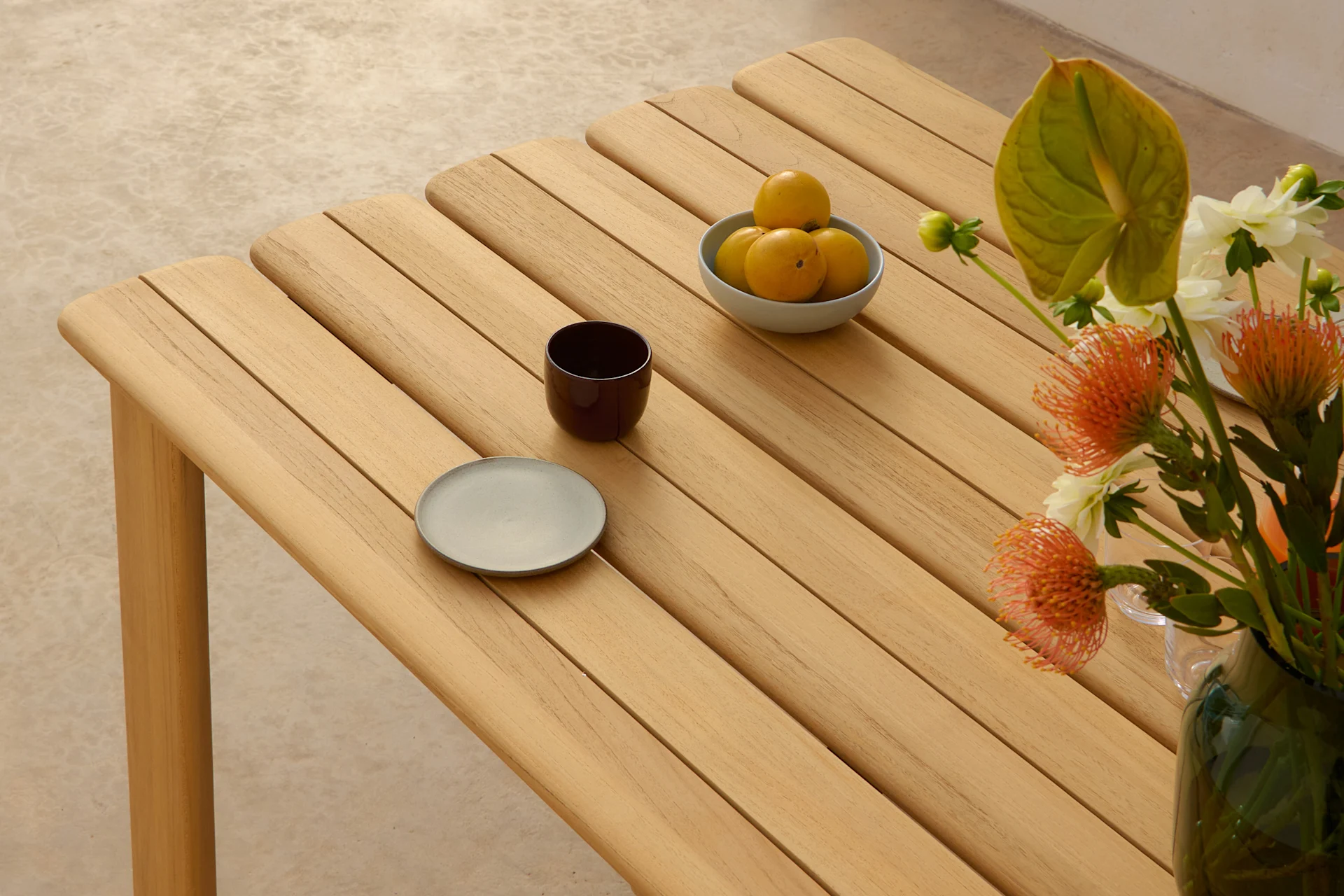 Una mesa de madera clara con listones, con un cuenco de frutas amarillas, una taza oscura, un plato gris y un jarrón de flores naranjas y blancas.