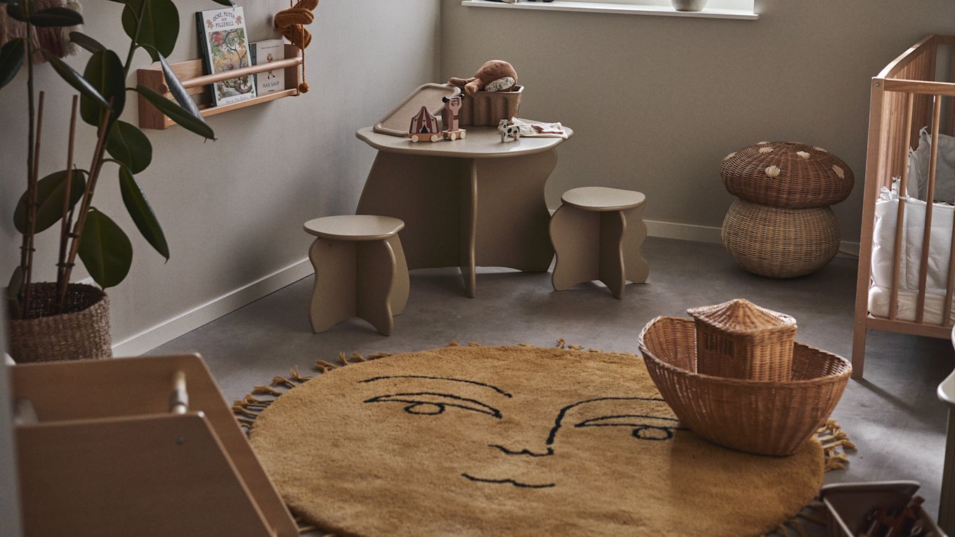 Colocar una alfombra grande y suave en el centro de la habitación crea una zona de juego acogedora y amplia a la hora de decorar la habitación infantil. Aquí la alfombra con forma de sol Sun de Ferm Living en una habitación infantil con mesa, taburetes y cuna.