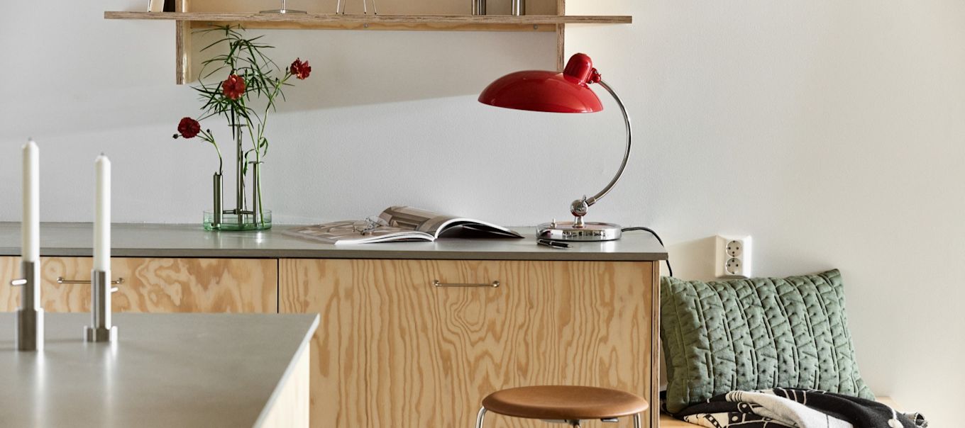 Aquí puedes ver la lámpara de mesa Kaiser en rojo rubí con el jarrón Ikeru en acero cepillado, ambos de Fritz Hansen, en una moderna cocina de madera contrachapada.