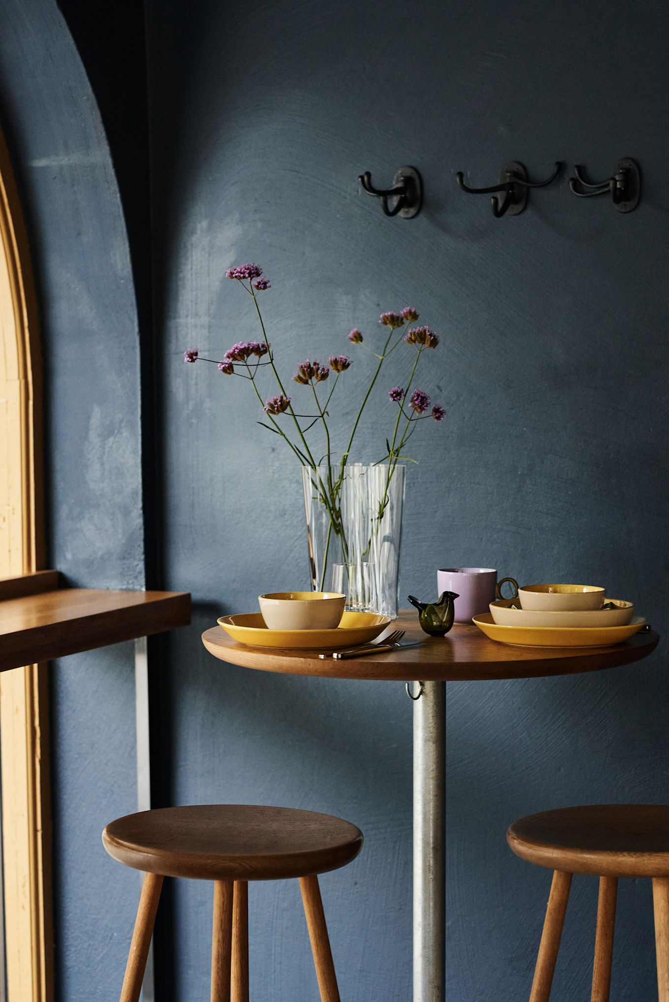 Aquí puedes ver una decoración de mesa retro en amarillo y púrpura en una habitación de color azul oscuro, con un jarrón brillante y flores púrpuras a juego.