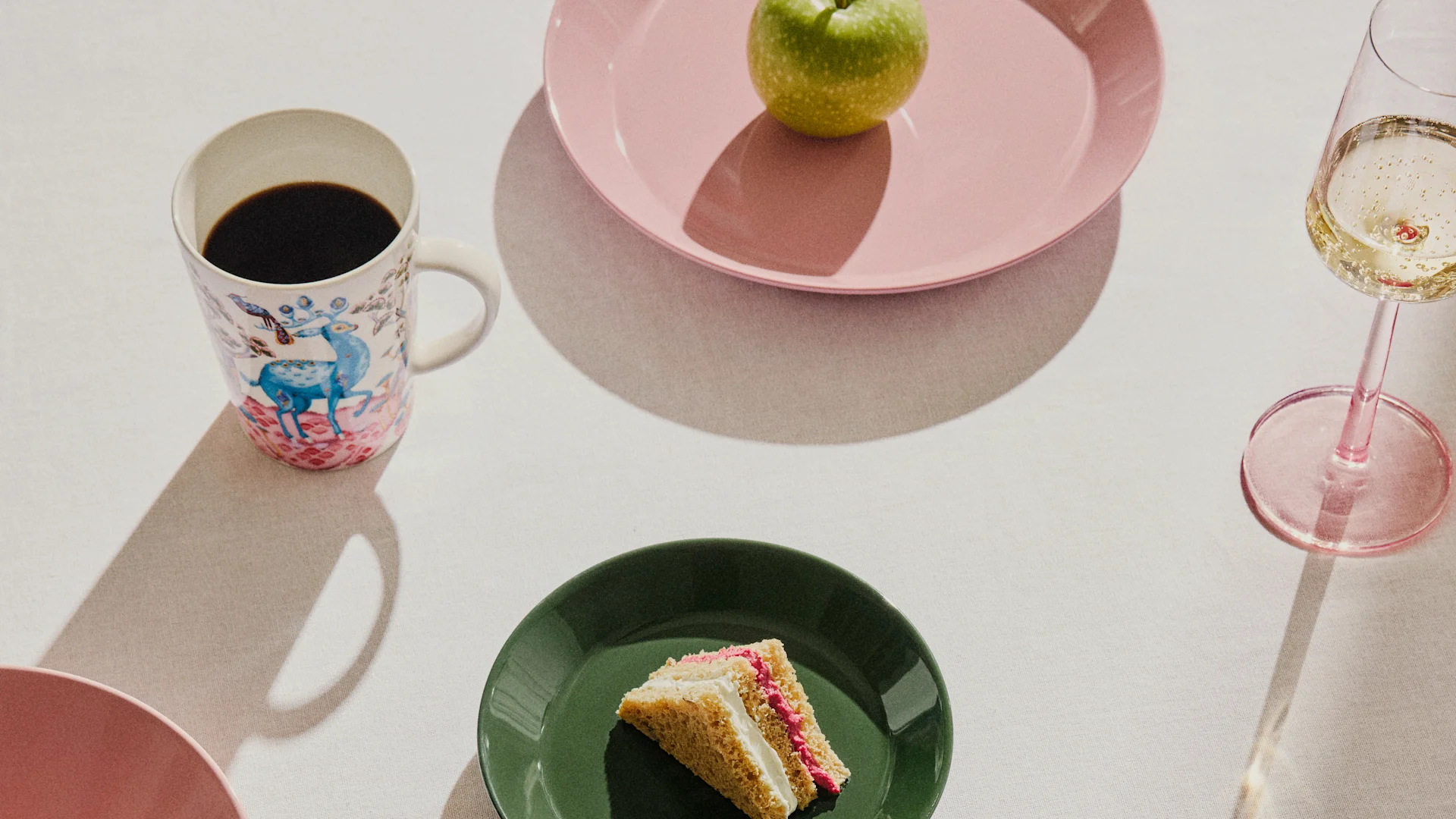 Mesa puesta con una taza de café estampada, una manzana verde en un plato rosa, un trozo de pastel en un plato verde y una copa de champán.