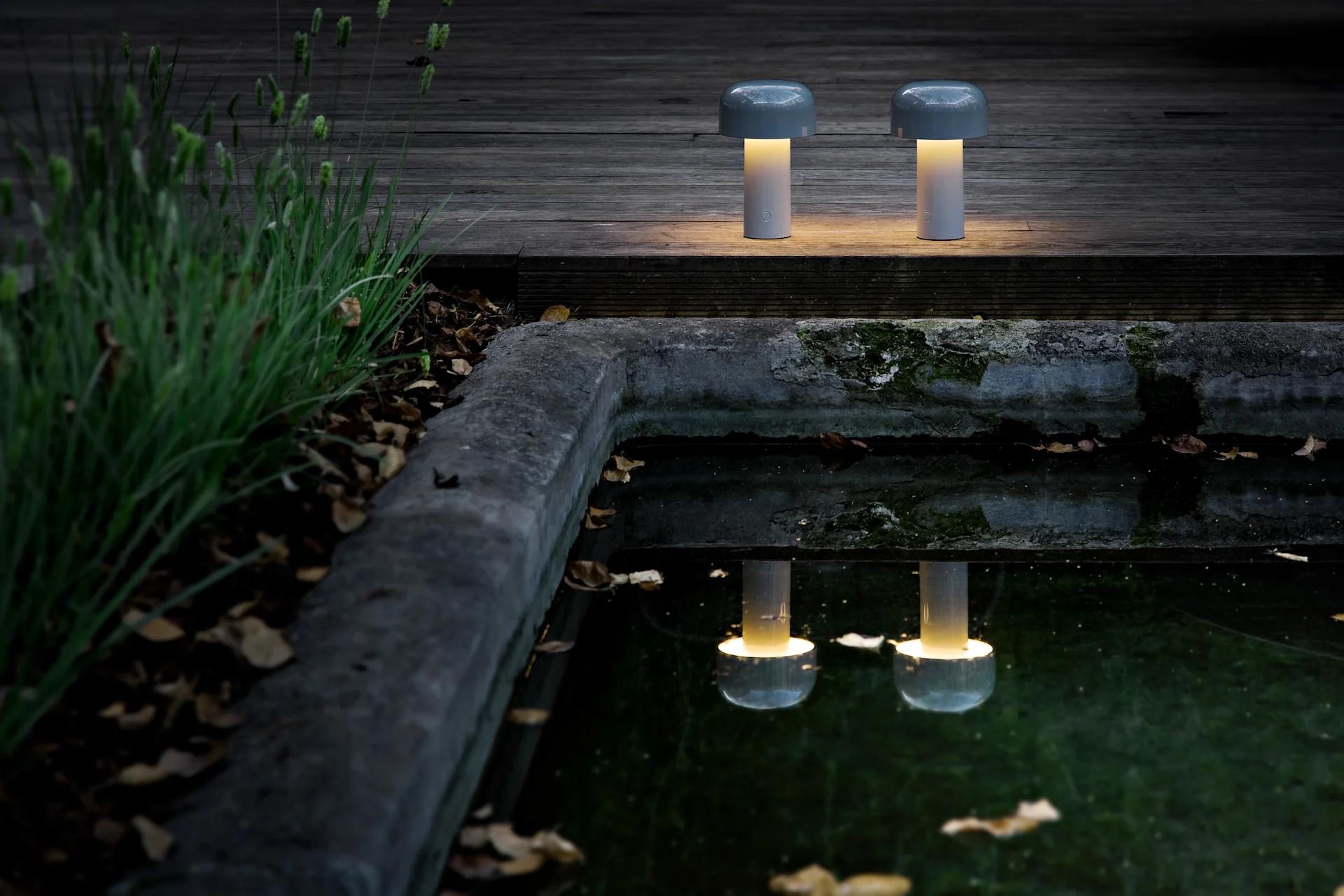 Dos lámparas de mesa portátiles en forma de hongo con luz amarilla cálida sobre una cubierta de madera oscura junto a un estanque, reflejando su resplandor.
