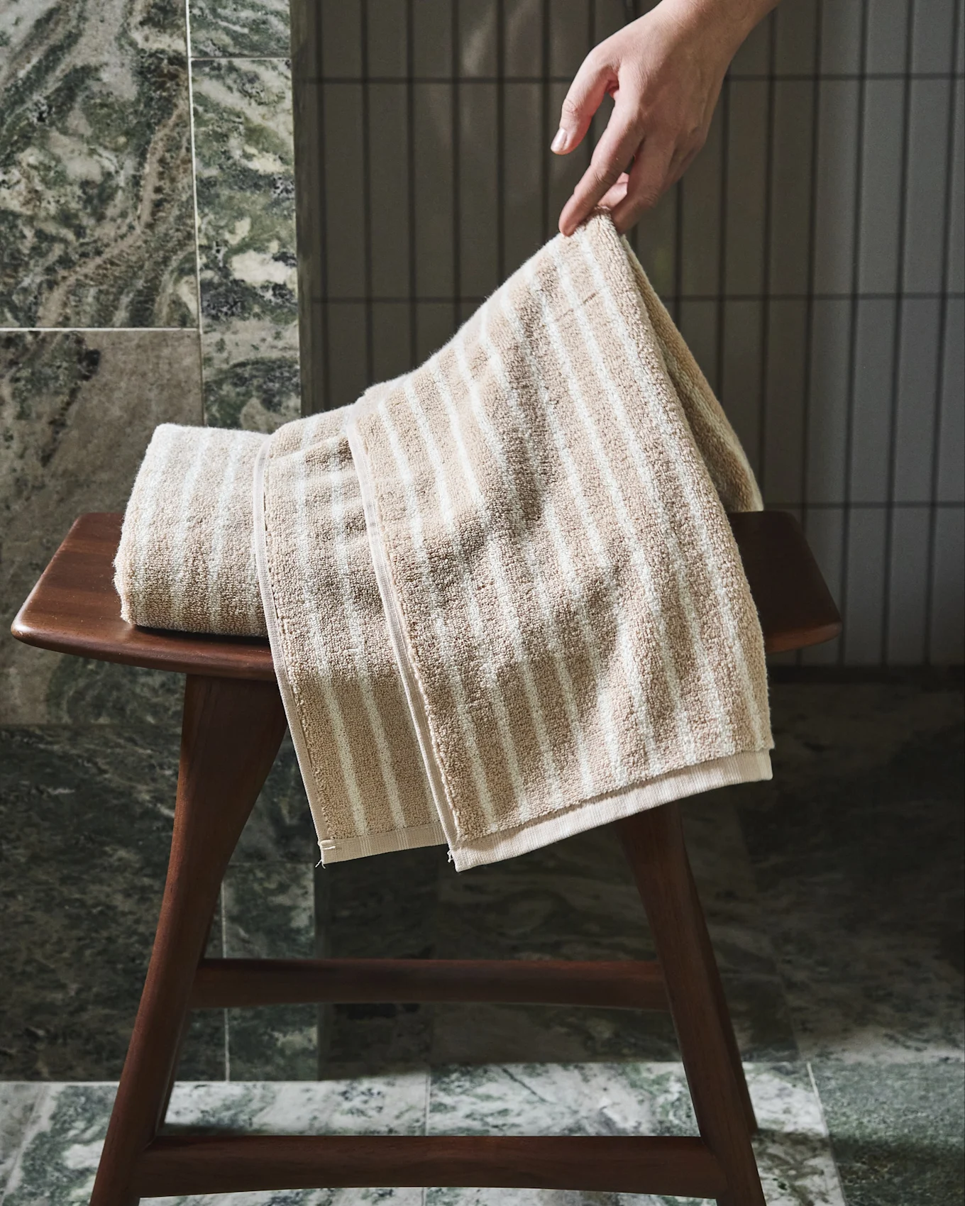 Mano ajustando una toalla a rayas beige y blanca sobre un taburete de madera.
