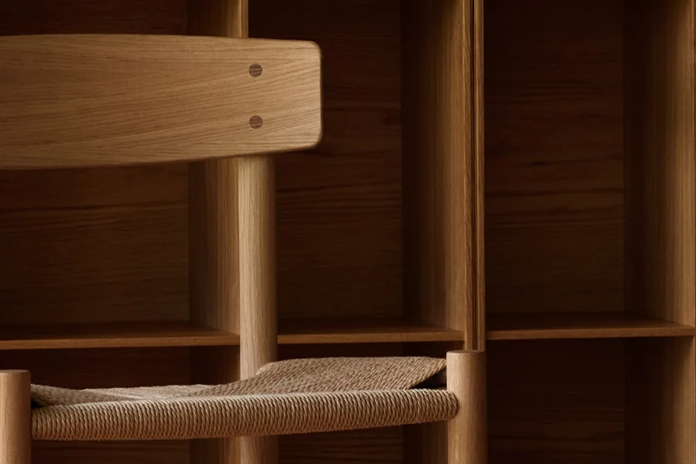 Una silla de madera clara con asiento trenzado frente a una pared de estantes cuadrados de madera y un suelo de madera con diseño.