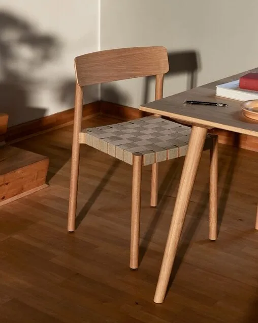 Una silla de madera clara con asiento de mimbre a cuadros junto a una mesa con libros y un bolígrafo.