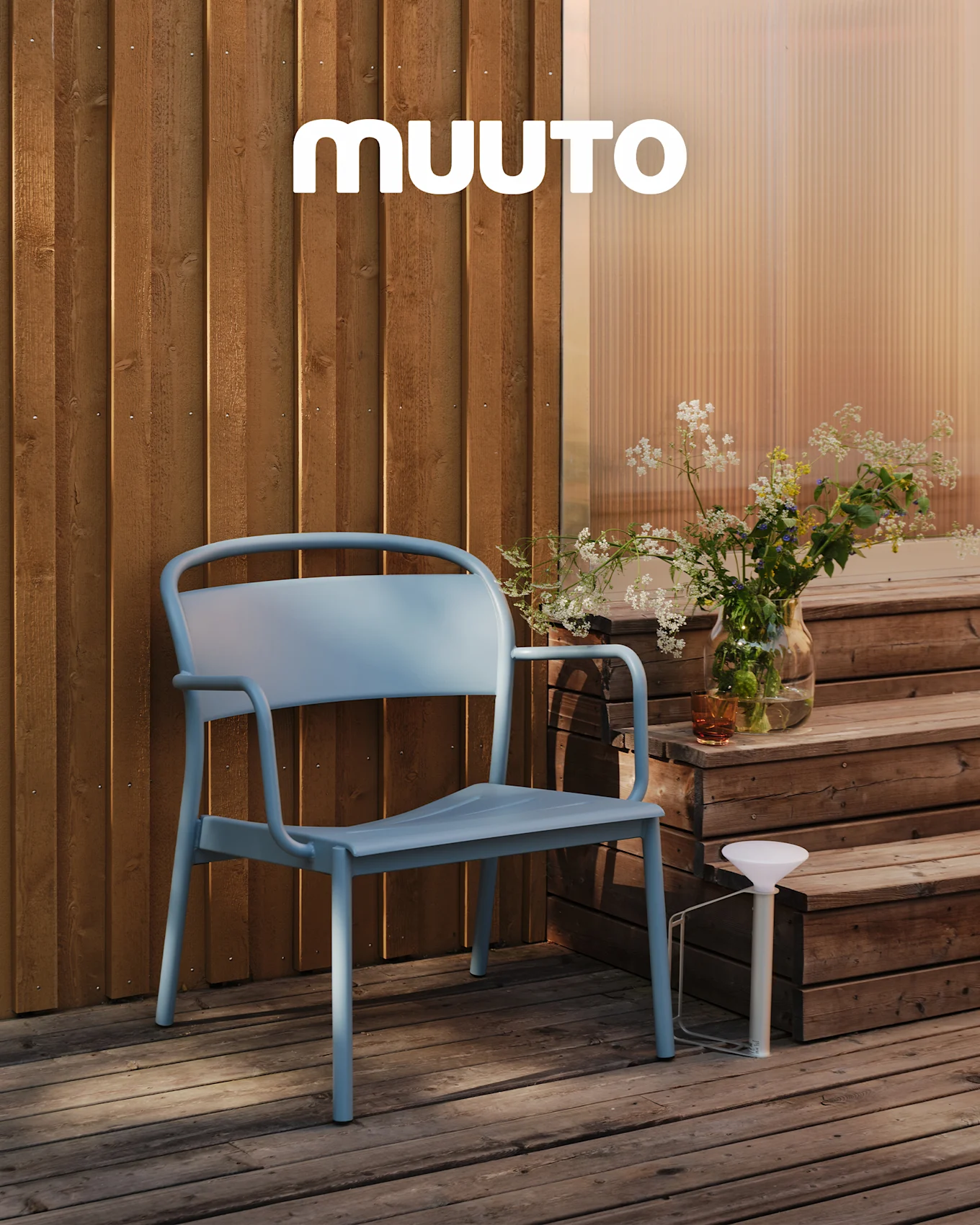 Una silla exterior azul claro en una terraza de madera junto a escalones con un jarrón de flores silvestres y una lámpara portátil blanca. Logo MUUTO.