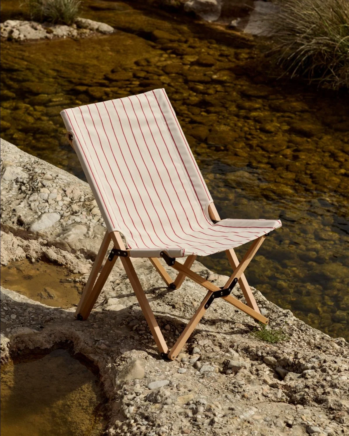 Silla plegable con asiento de tela a rayas blancas y rojas en una orilla rocosa junto a un río.