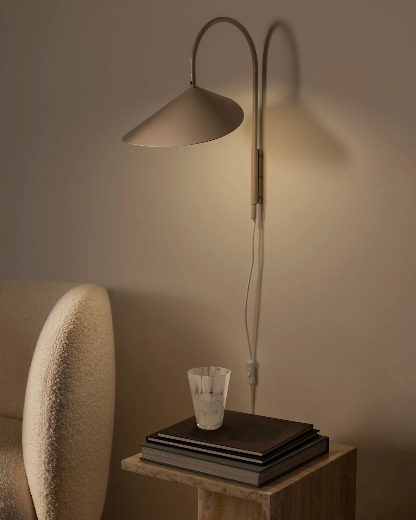 Lámpara de pared cálidamente iluminada con pantalla cónica sobre pila de libros y vaso texturizado en mesa de piedra, junto a sillón beige.
