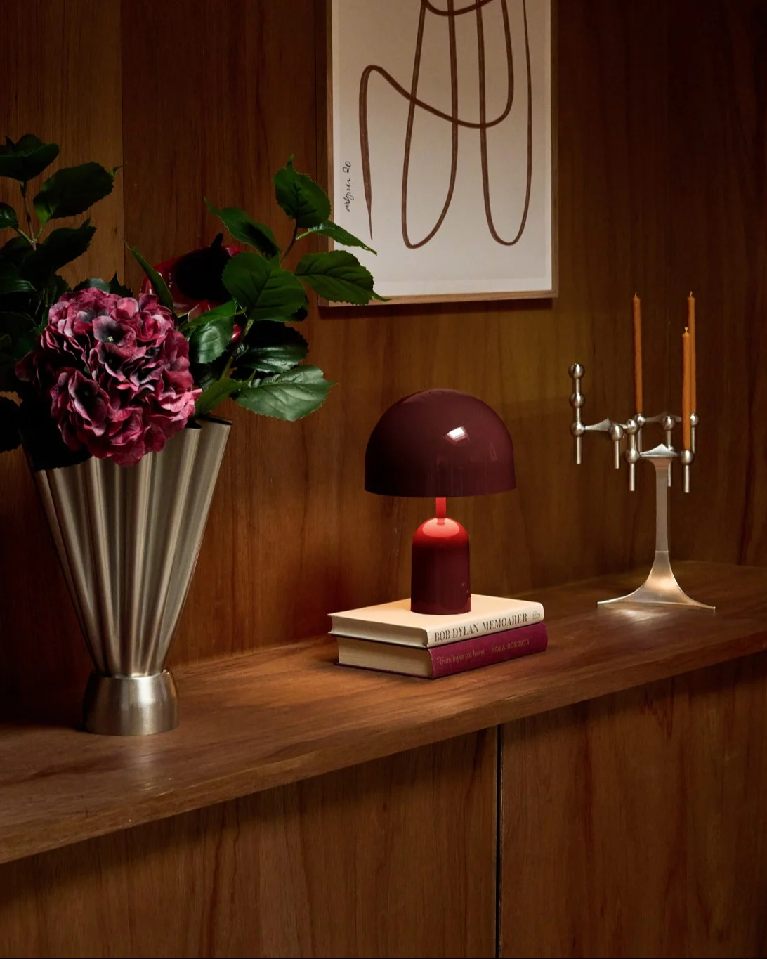 Acogedora estantería de madera con jarrón plateado con hortensias rojas, lámpara roja sobre libros y candelabro plateado.