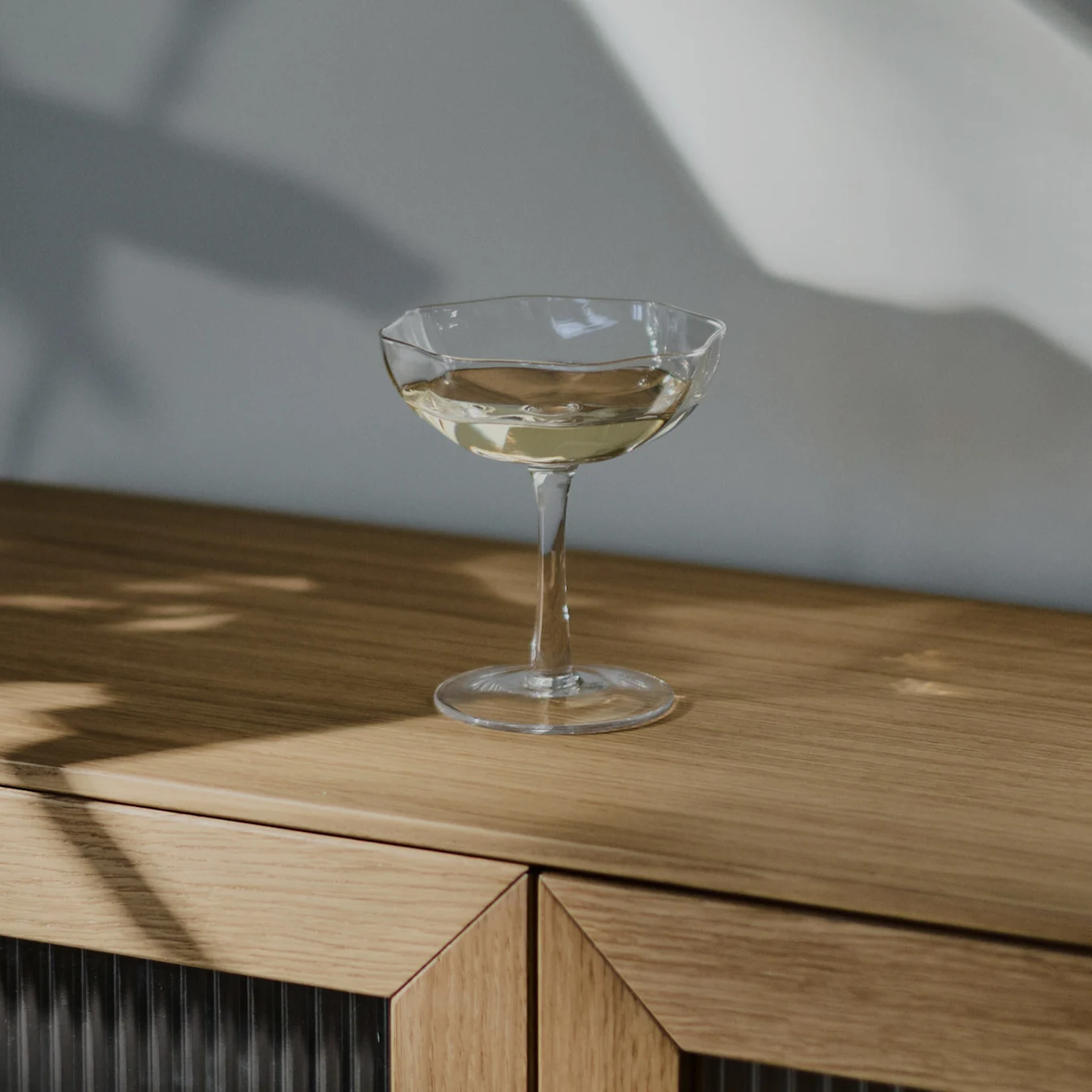 Una copa de cóctel transparente con una bebida de color claro sobre un mueble de madera clara, con sombras en la pared.