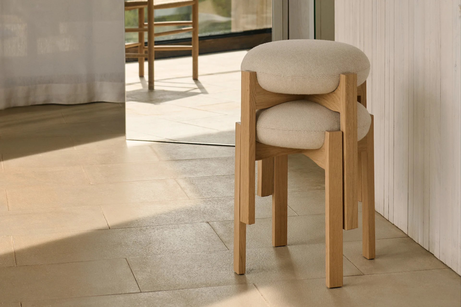 Dos taburetes minimalistas de madera clara con asientos tapizados redondos apilados sobre baldosas claras, con vista soleada al exterior a través de una ventana grande.