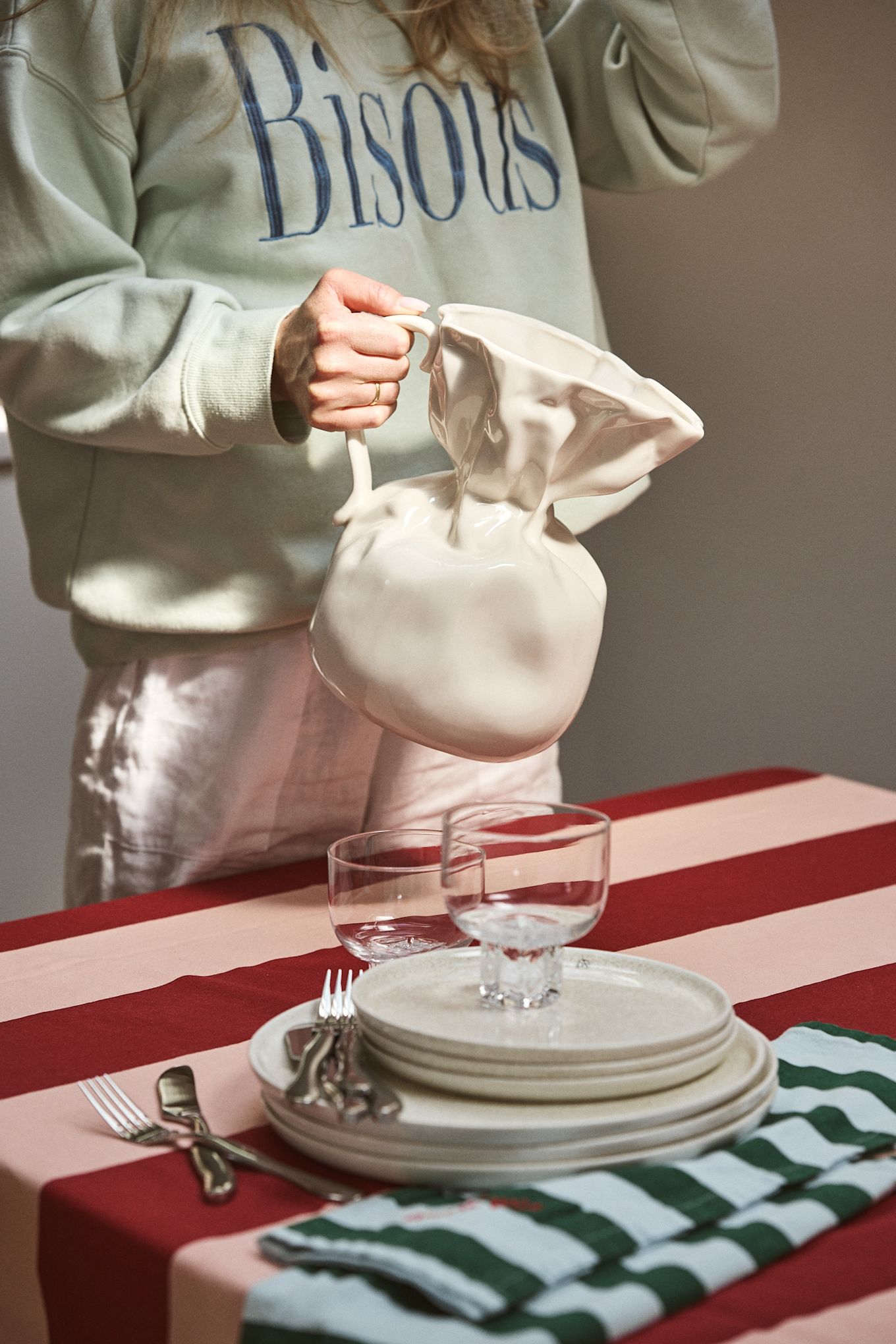 Persona sosteniendo un jarrón Crumple en blanco de Byon en una mesa puesta con mantel a rayas en rosa y burdeos. El jarrón arrugado tiene forma de bolsa de papel doblada, realizado en cerámica esmaltada.