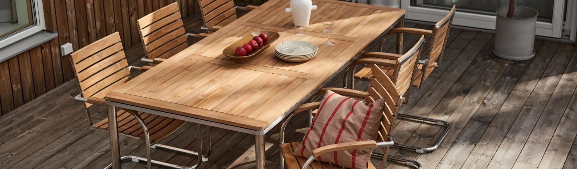 Mesa de comedor de madera para exterior con varias sillas en una terraza. La mesa tiene manzanas, platos y vasos. Una silla tiene un cojín a rayas.