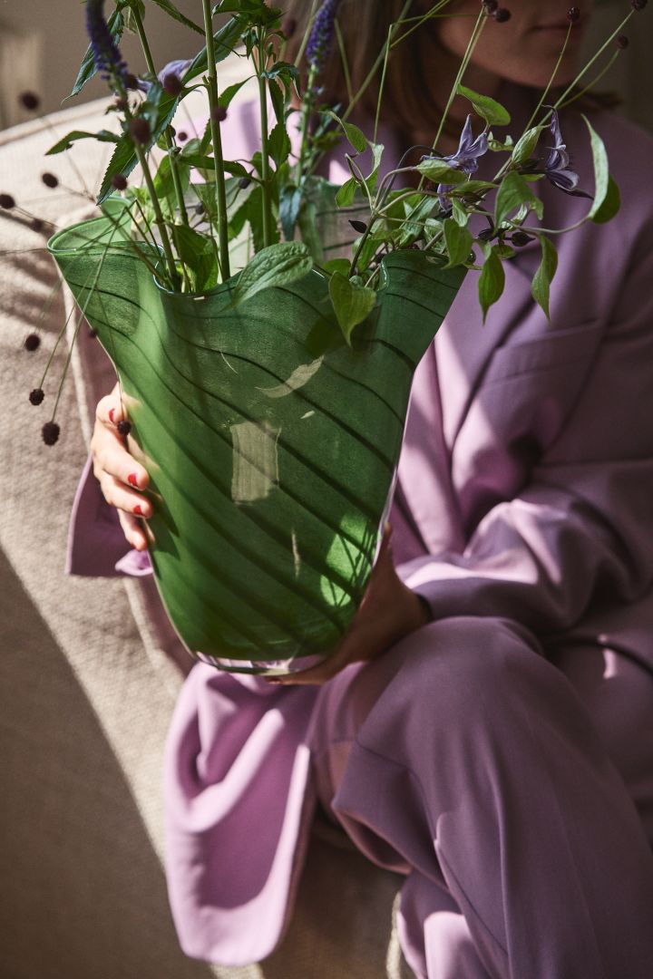 Persona vestida de morado sosteniendo el jarrón de cristal verde Tiggy de Byon, un jarrón plisado con forma de campana en verde guisante con rayas.