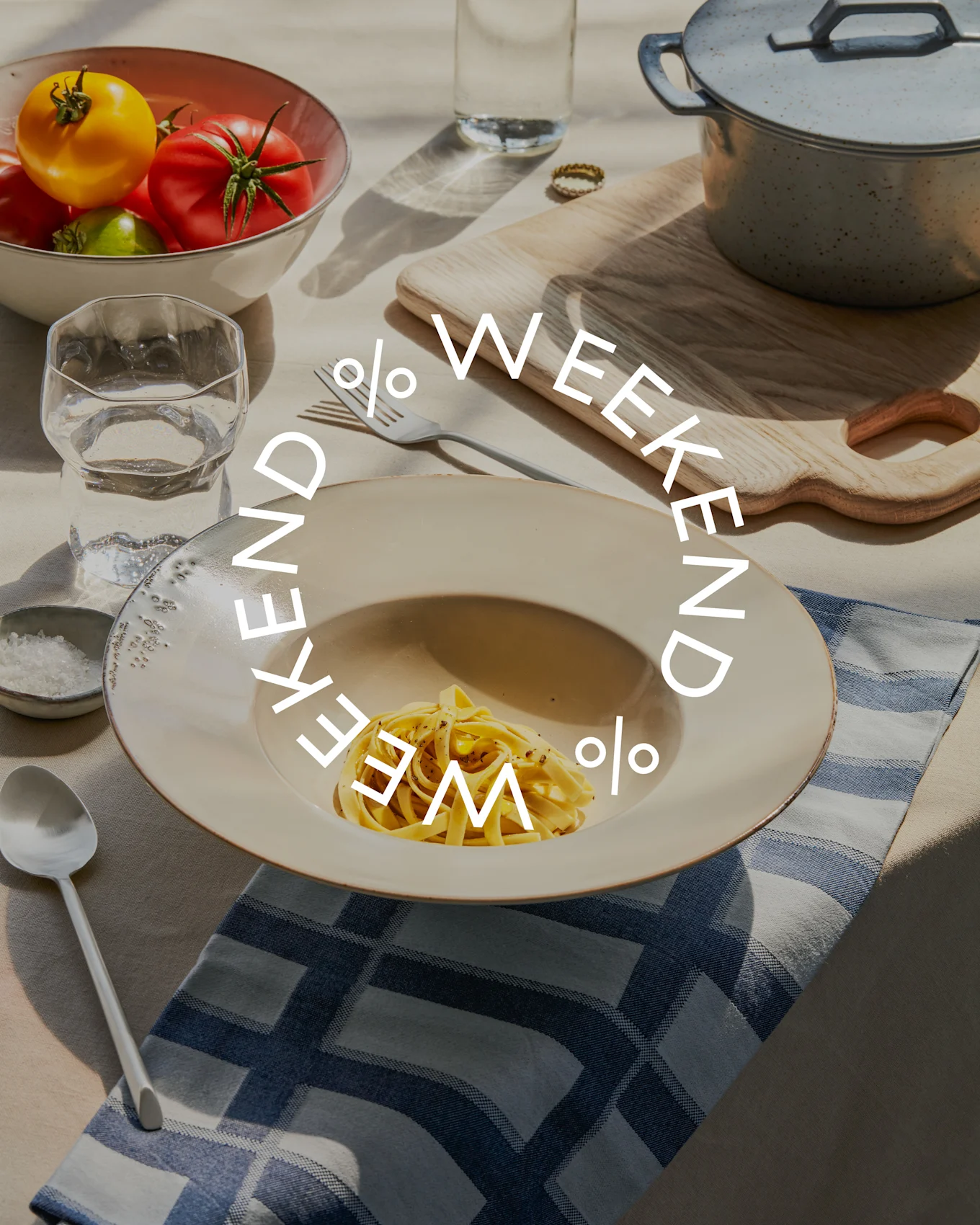 Mesa con pasta, un cuenco de tomates frescos, vaso de agua y olla de cocción. El texto 'WEEKEND % WEEKEND %' está superpuesto.