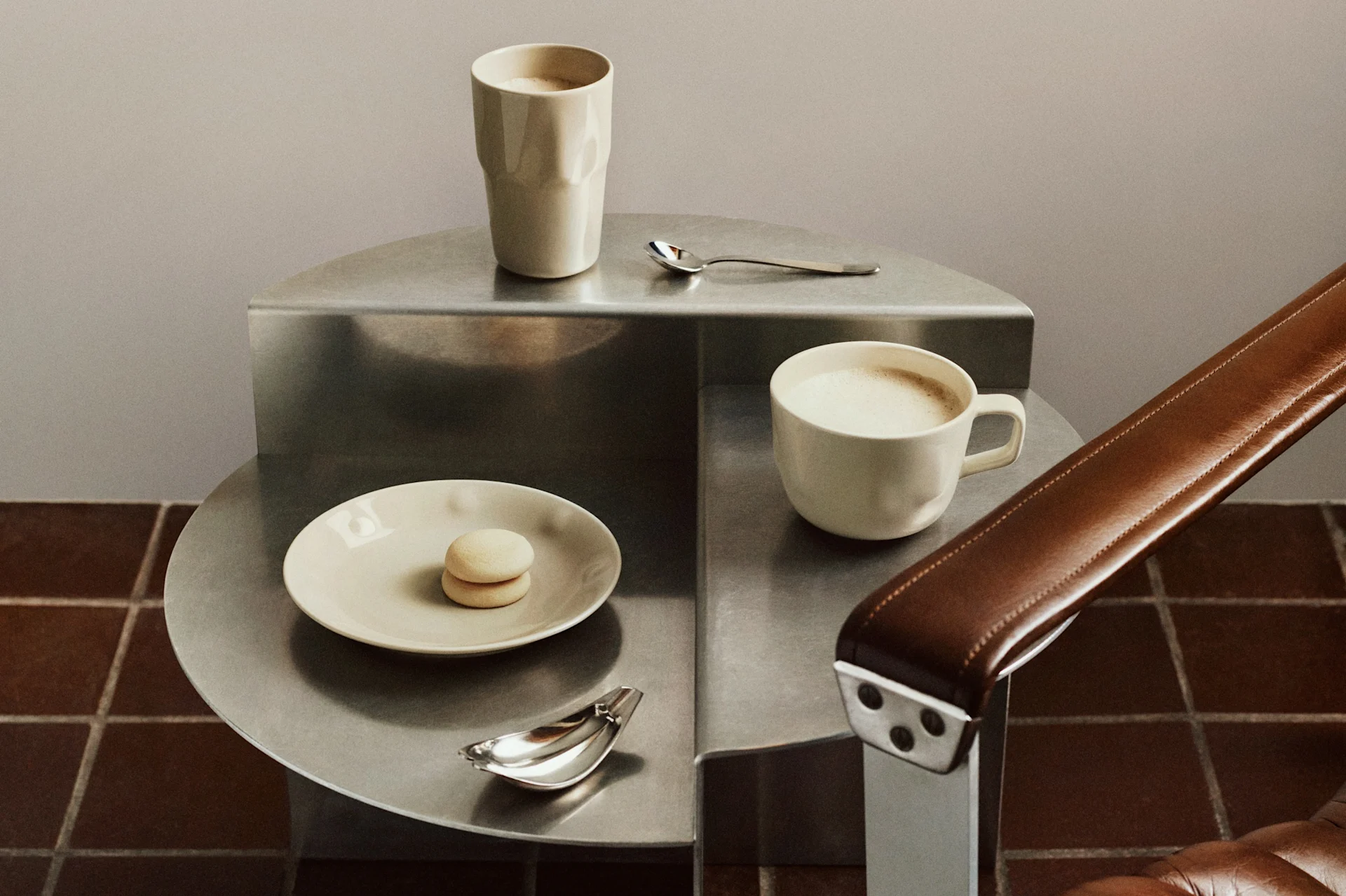 Dos tazas de café claras, un macaron y cucharas en una mesa auxiliar metálica escalonada junto a un sillón de cuero marrón.