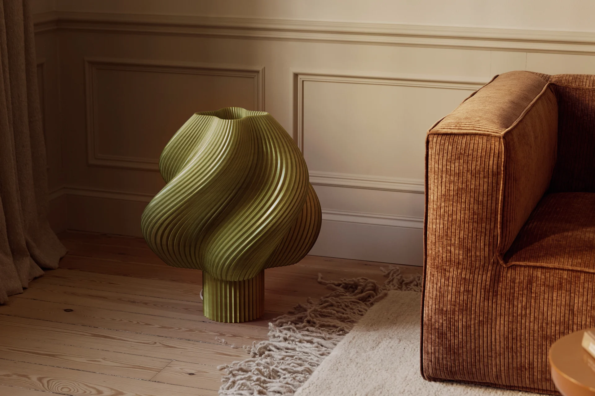 Un objeto escultural verde oliva estriado sobre un suelo de madera junto a un sofá de pana marrón y una alfombra texturizada en una habitación con paredes moldeadas.