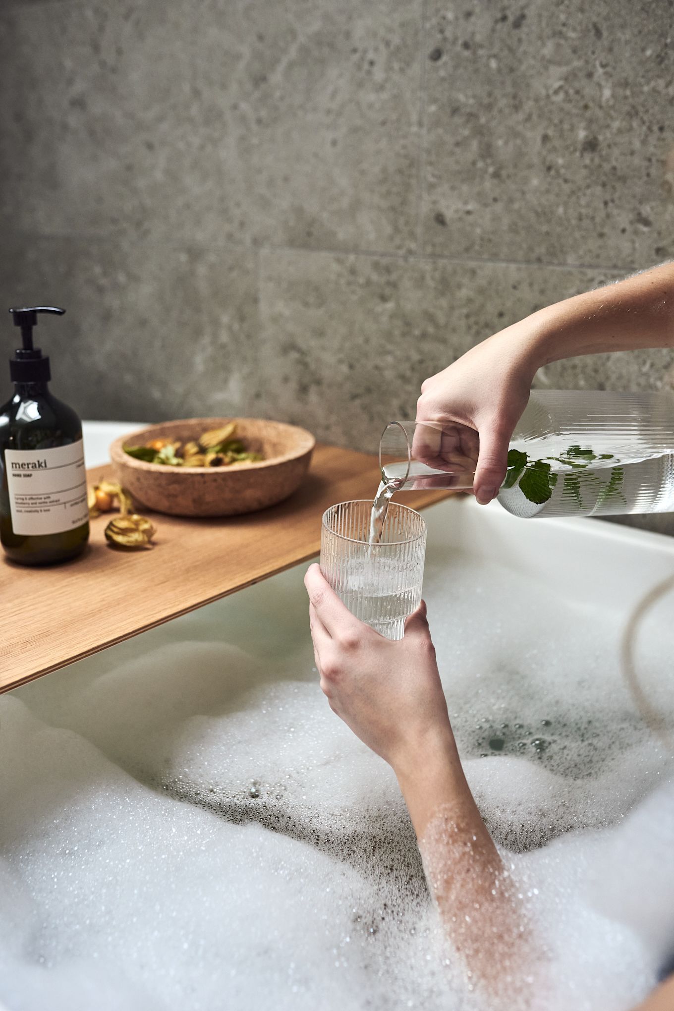 Unas manos vertiendo agua en un vaso ondulado de Ferm Living como parte de una noche de spa en casa con un cálido baño de burbujas.