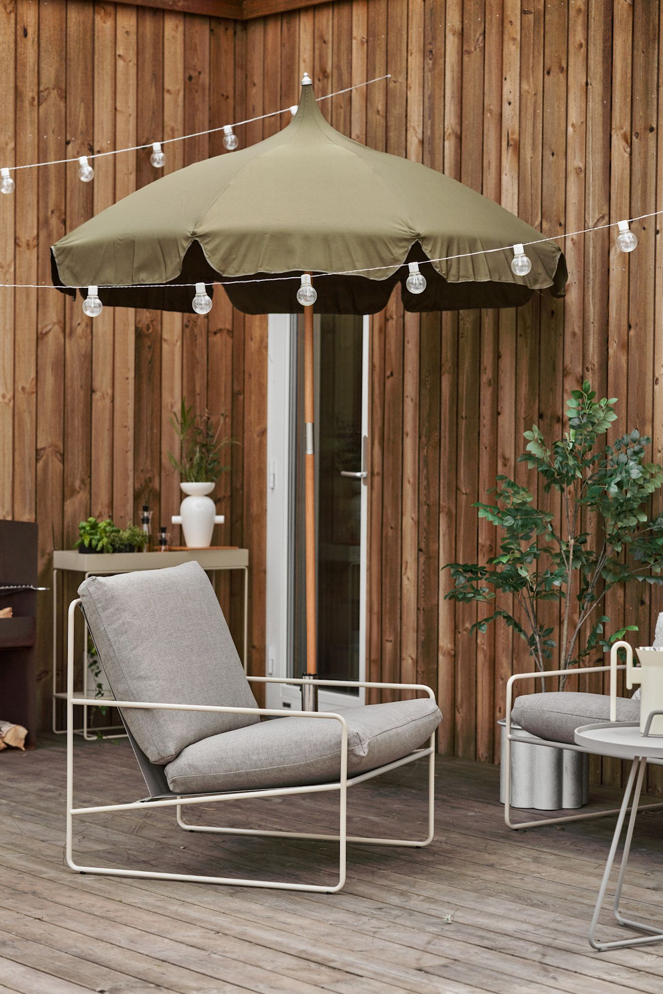 Una terraza de madera acogedora con guirnalda de luces, sombrilla Lull y silla lounge Desert de Ferm Living.