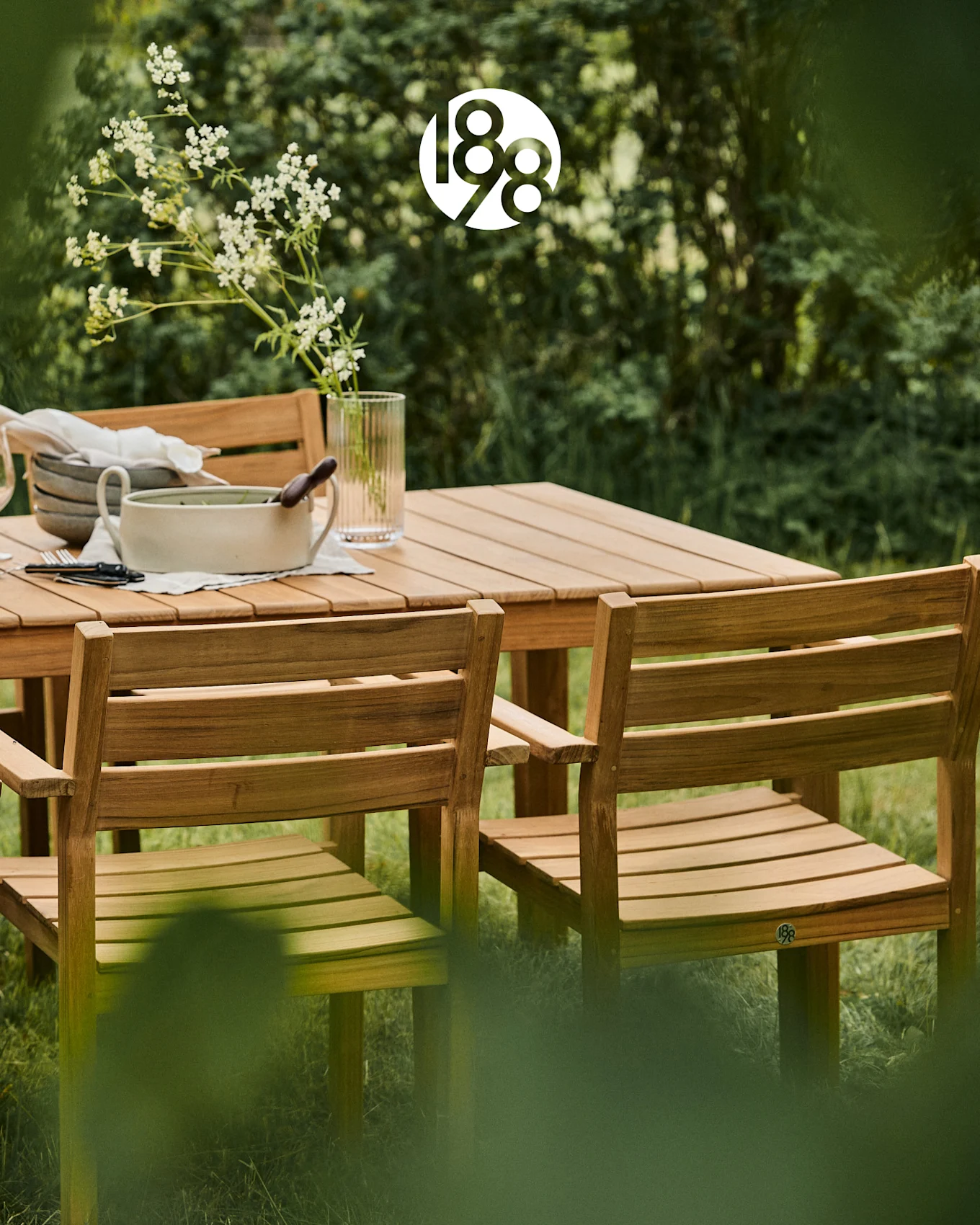 Conjunto de comedor de madera para exterior con mesa y sillas en un exuberante jardín verde, con flores blancas y vajilla.
