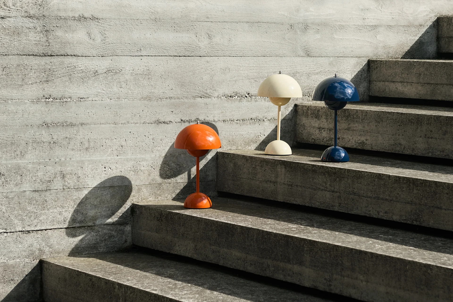 Tres lámparas de mesa con forma de cúpula en naranja, crema y azul sobre escaleras de hormigón junto a una pared de hormigón, proyectando sombras.