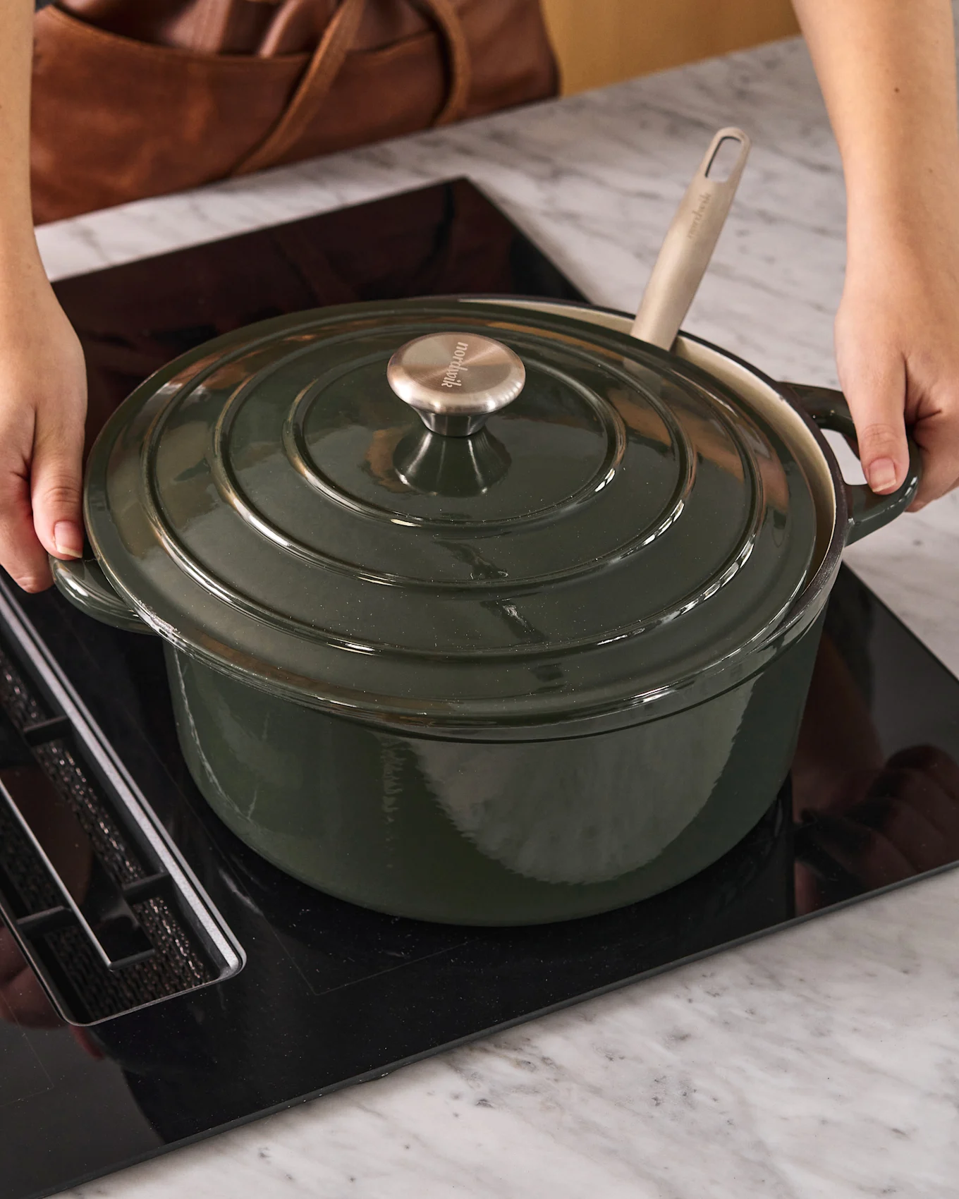 Persona colocando una olla de hierro fundido verde oscuro con pomo metálico sobre una placa de inducción negra.